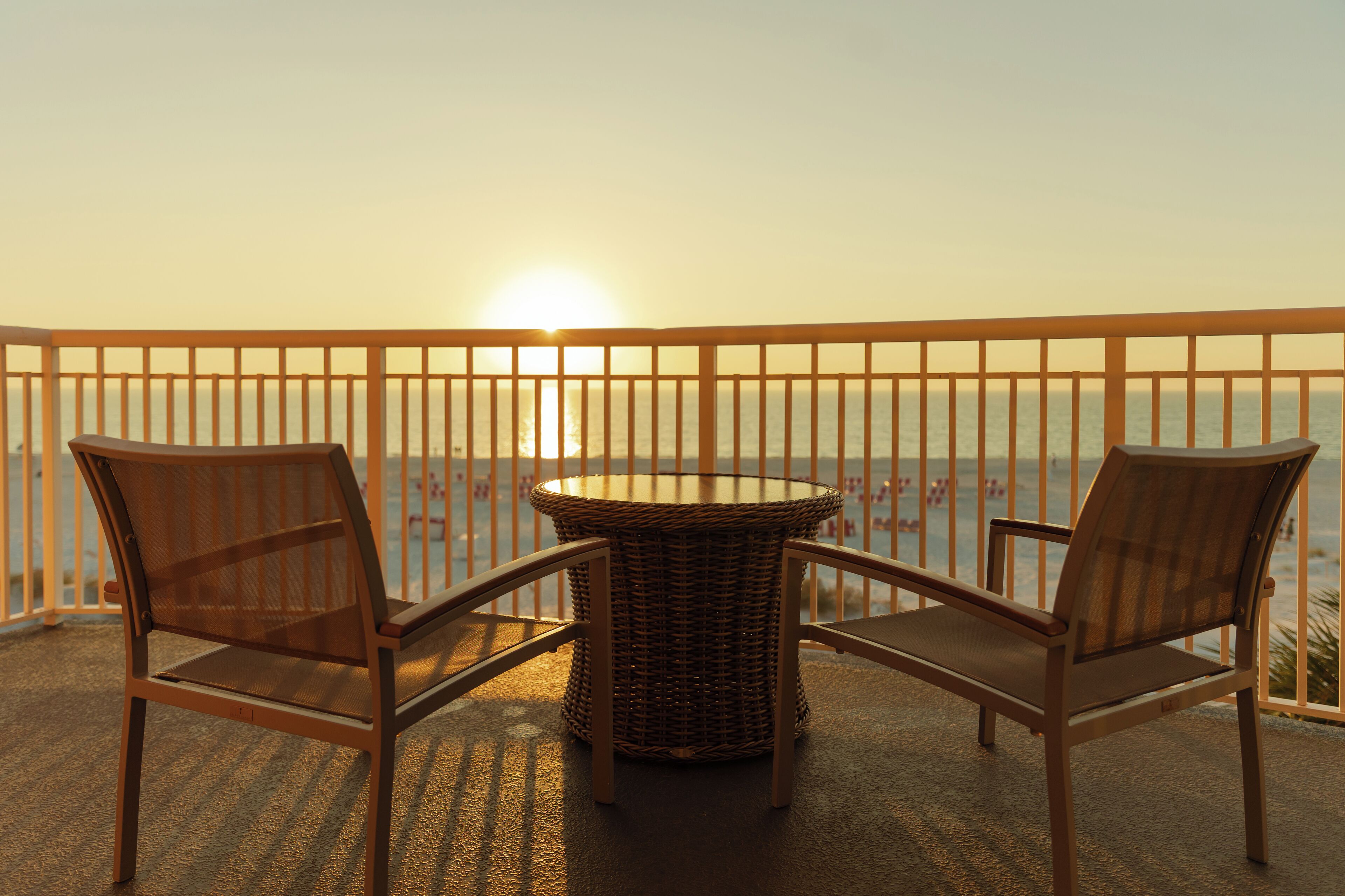 A balcony with two wicker chairs and a small table, overlooking a body of water during sunset.