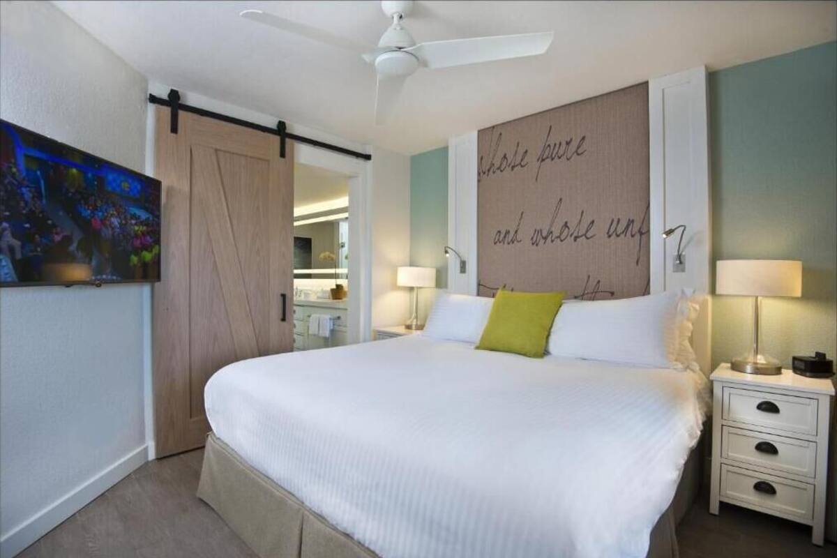 A bedroom with a bed, bedside tables, a ceiling fan, a wall with a motivational quote, and a TV on the wall.