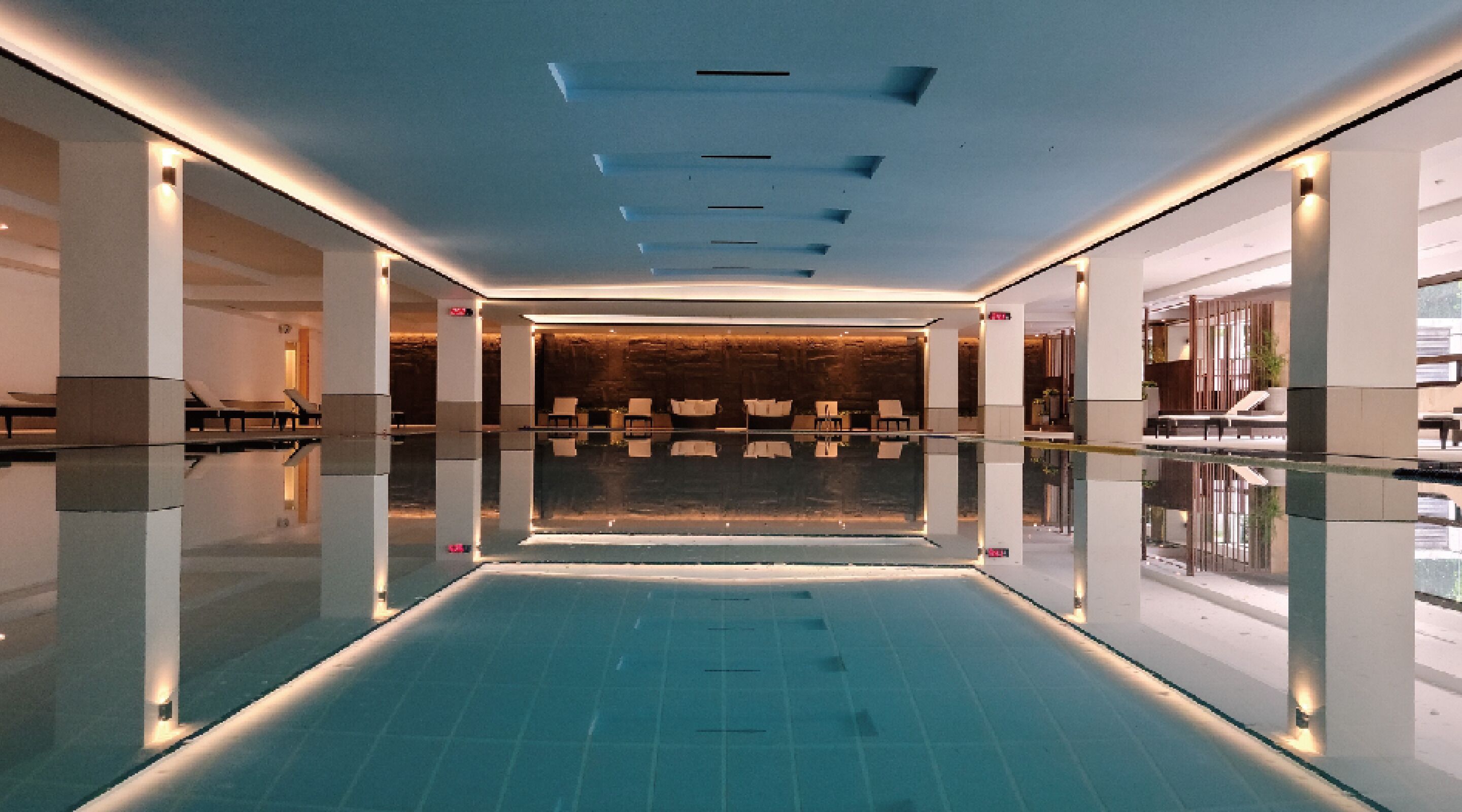 A modern indoor swimming pool with a clear view of the pool's edge, a seating area with chairs, and a reception desk in the background.