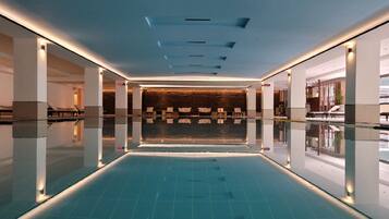 A modern indoor swimming pool with a clear view of the pool's edge, a seating area with chairs, and a reception desk in the background.