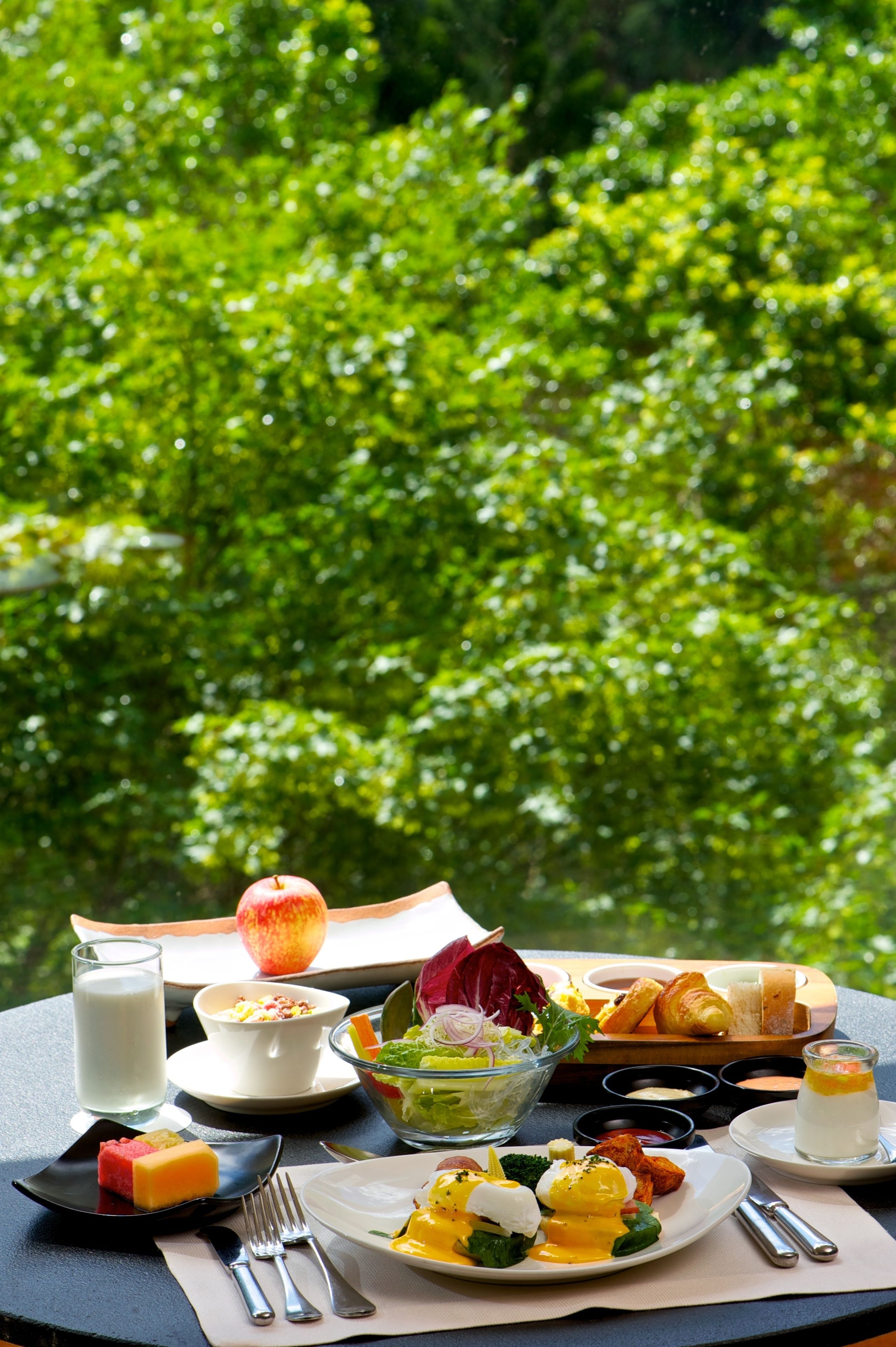 A table set with breakfast items including eggs, toast, fruit, and a salad, with a glass of milk and a croissant, set against a backdrop of lush green foliage.