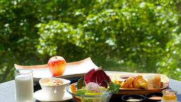 A table set with breakfast items including eggs, toast, fruit, and a salad, with a glass of milk and a croissant, set against a backdrop of lush green foliage.