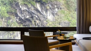 A room with a view of a rocky cliff and lush greenery. There is a wicker chair, a small round table with a vase of flowers, and a sofa. - Silks Place Taroko (Xiulin)