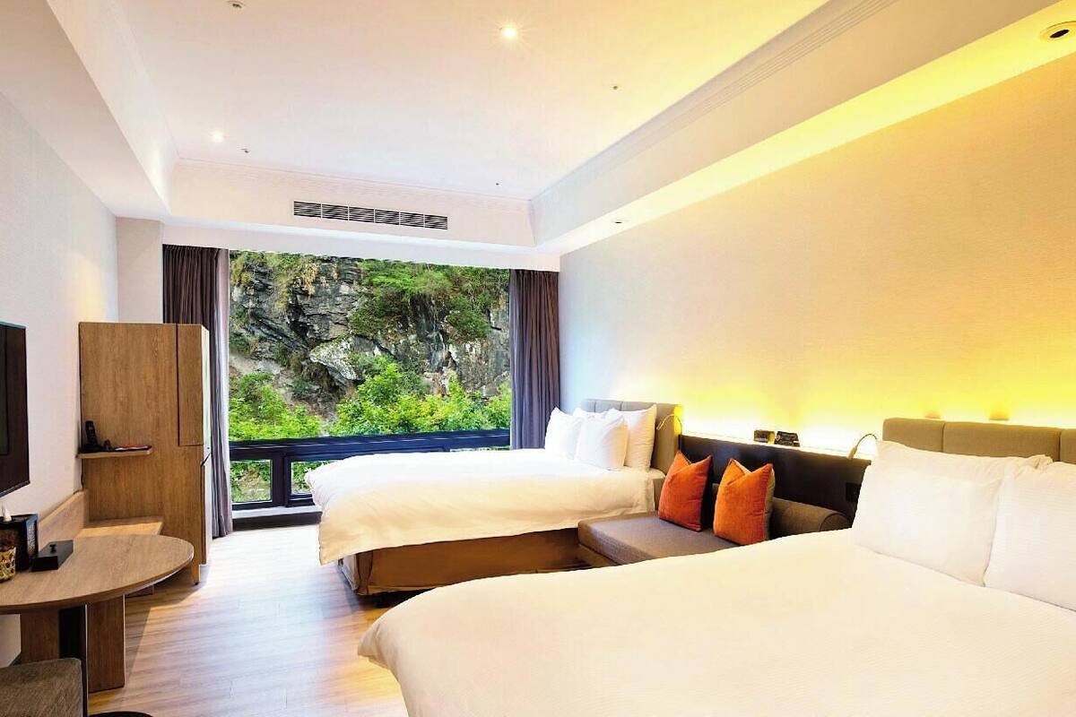 A hotel room with two beds, a TV, a desk, and a balcony with a view of greenery.
