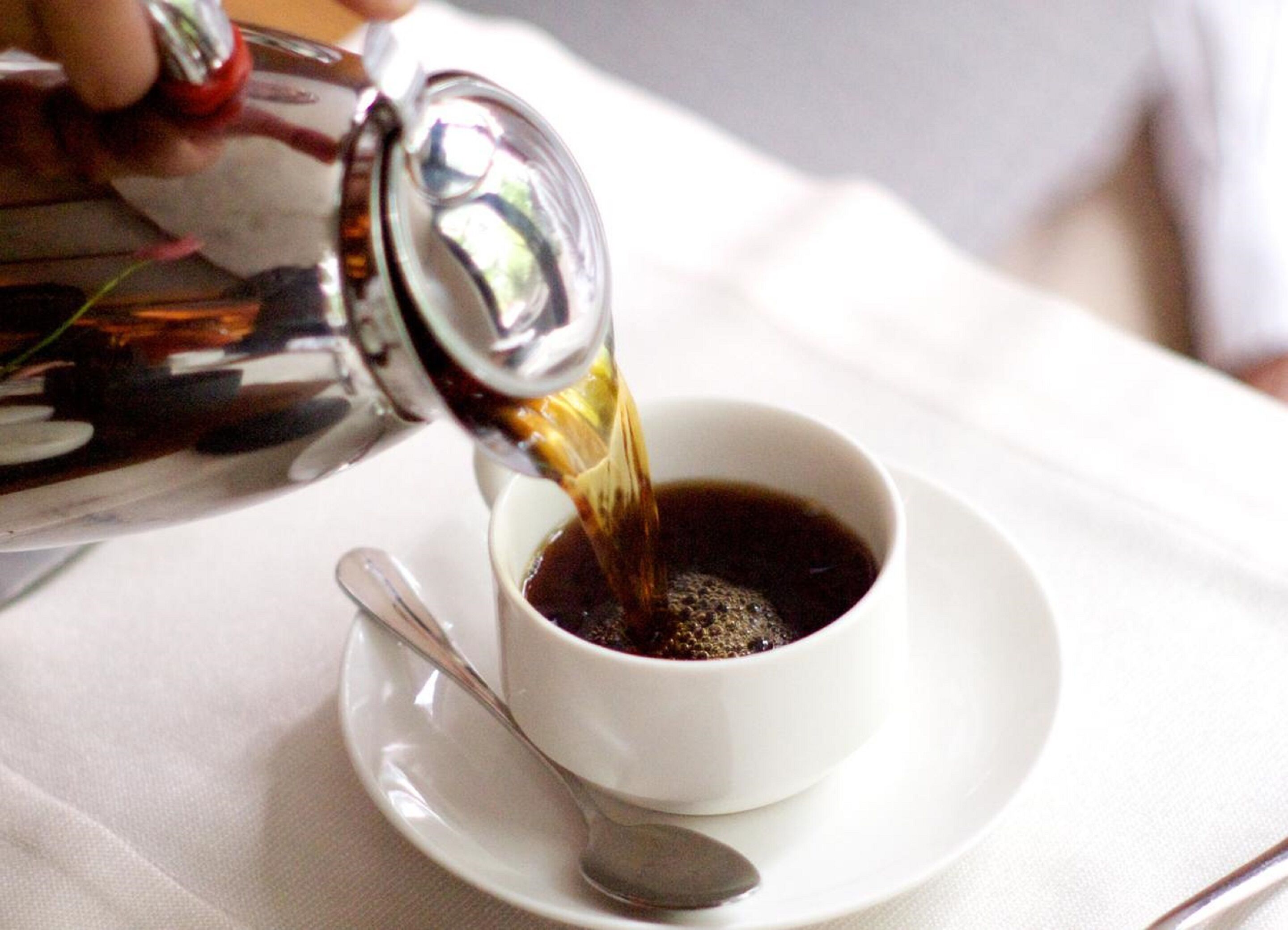 A cup of coffee being poured from a metallic pitcher into a white cup on a saucer.