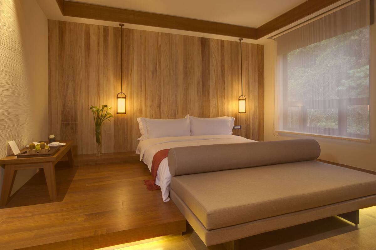 A modern hotel room with a wooden wall panel, a bed with white linens, a bedside table with a lamp, and a large window with a view of trees.