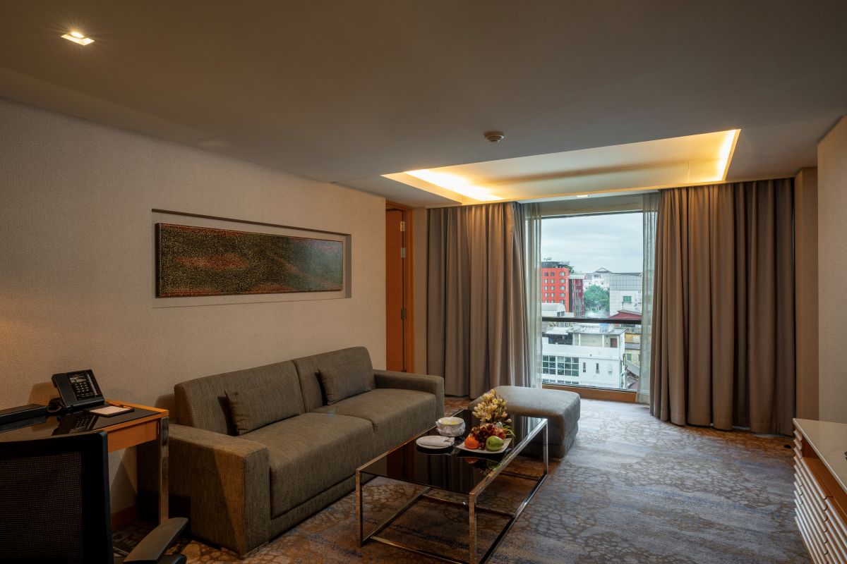 A hotel room with a sofa, a small table with a fruit plate, a painting on the wall, and a view of buildings through the window.