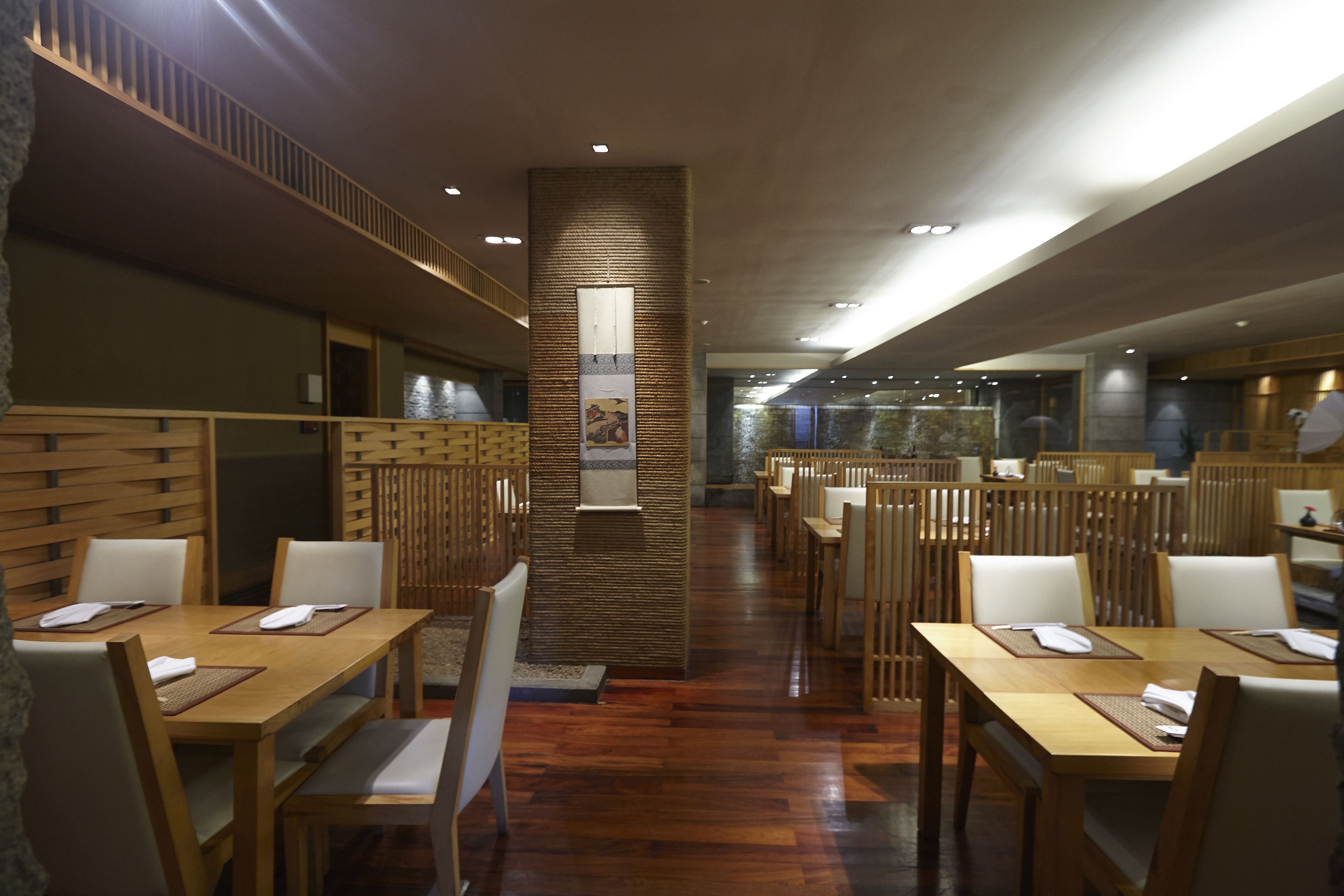 A restaurant with wooden tables and chairs, a central pillar with artwork, and a modern ceiling design.
