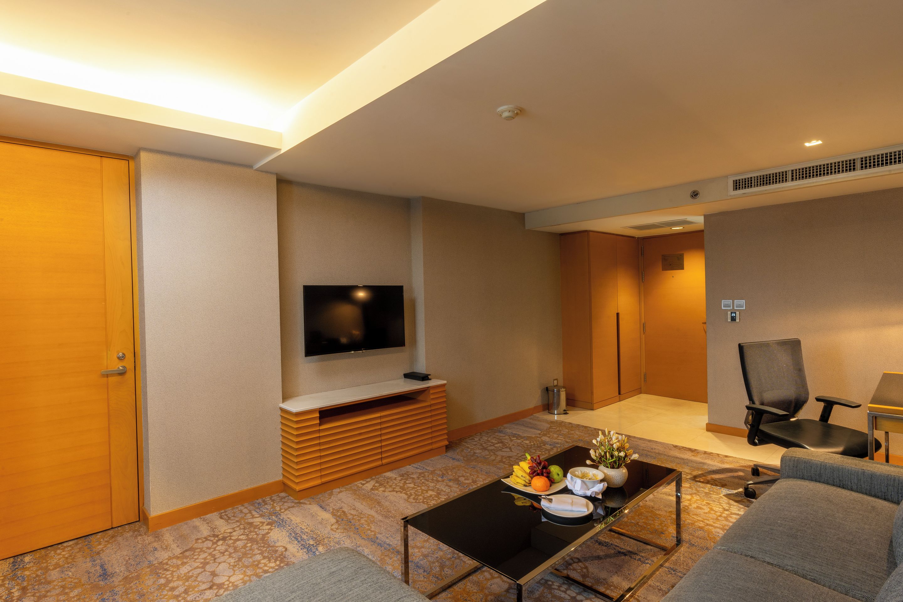 Habitación de hotel con televisión de pantalla plana en la pared, mueble de entretenimiento de madera, mesa de centro con frutas y un florero, un sofá y una silla.