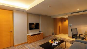 Habitación de hotel con televisión de pantalla plana en la pared, mueble de entretenimiento de madera, mesa de centro con frutas y un florero, un sofá y una silla.