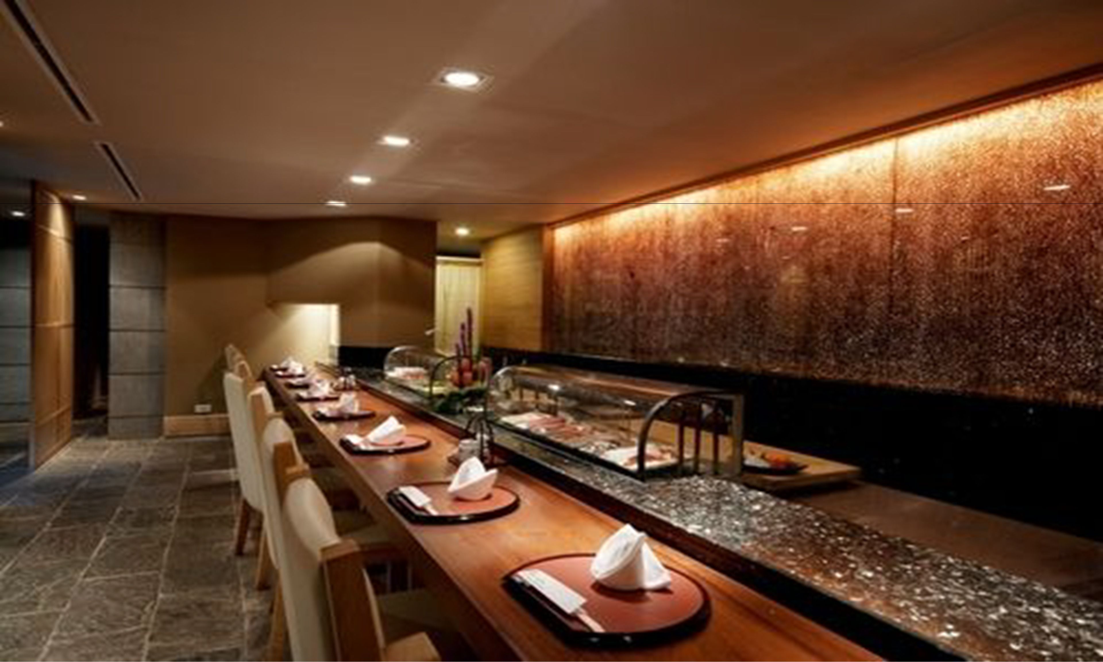 A long, wooden buffet table with neatly arranged plates and napkins, a glass display case with food, and a textured wall behind.