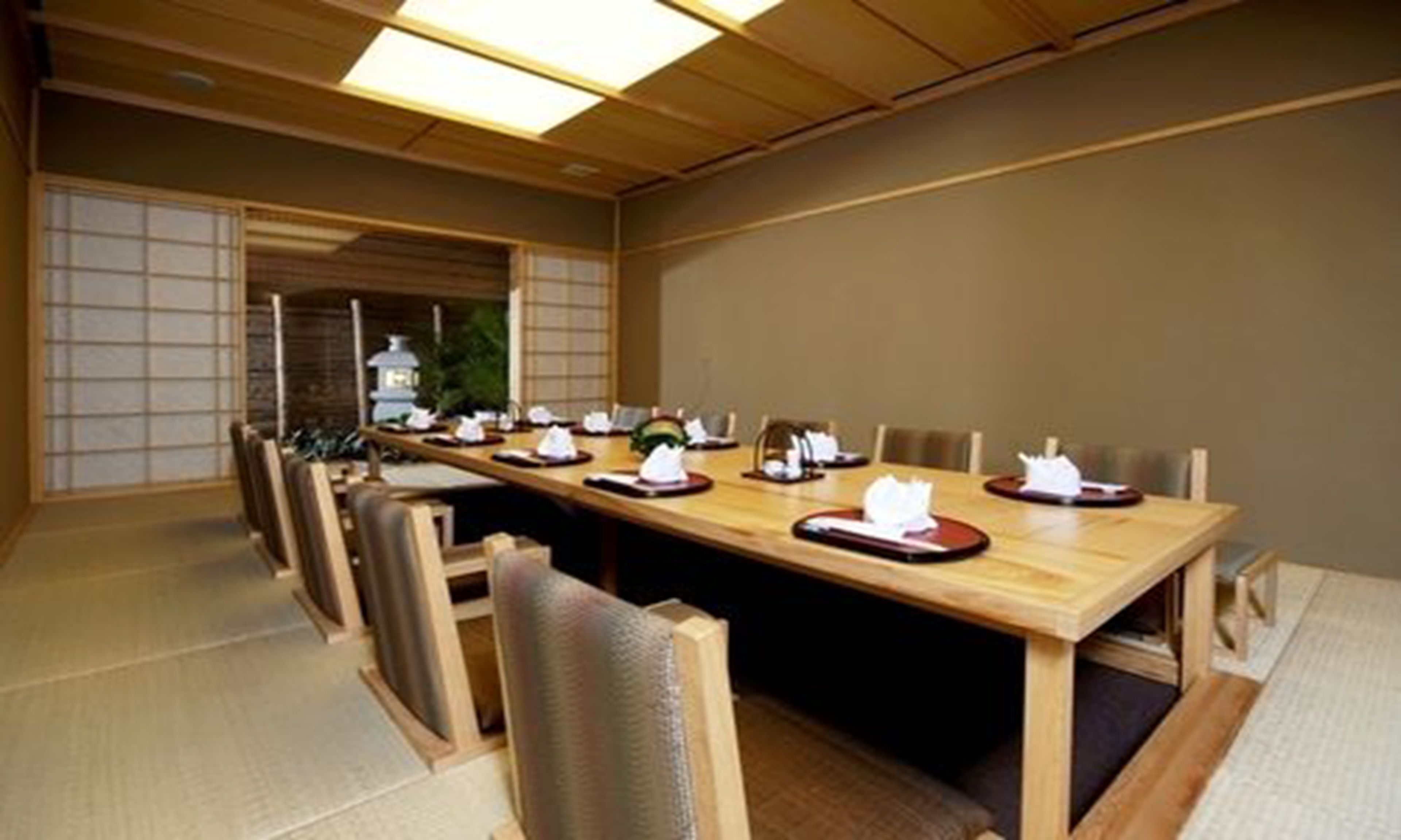 A traditional Japanese-style dining room with a long wooden table set for a meal, surrounded by tatami mat flooring and low wooden chairs.