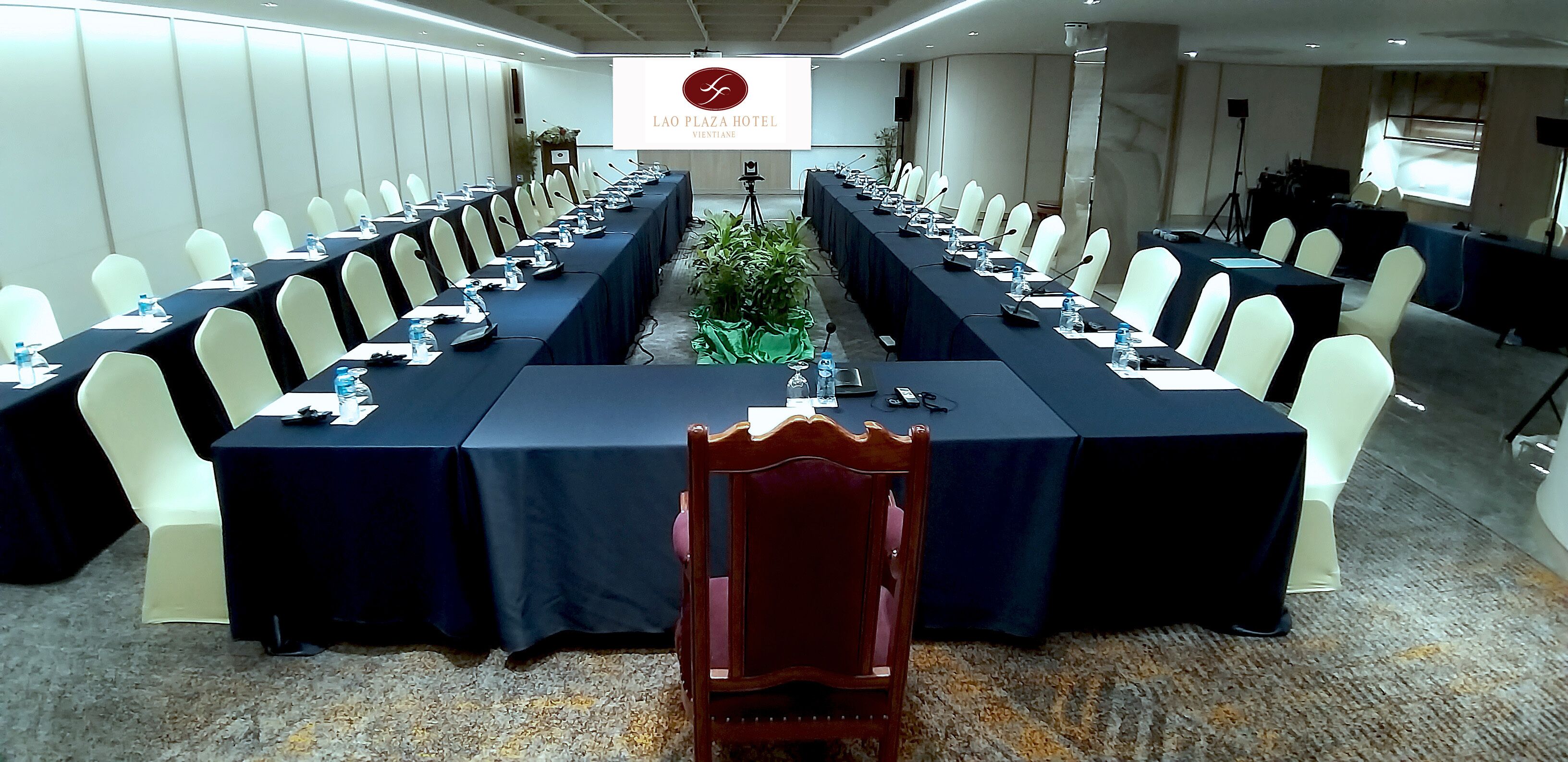 Un salón de conferencias con una larga mesa, sillas blancas y una pantalla que muestra el nombre del hotel.