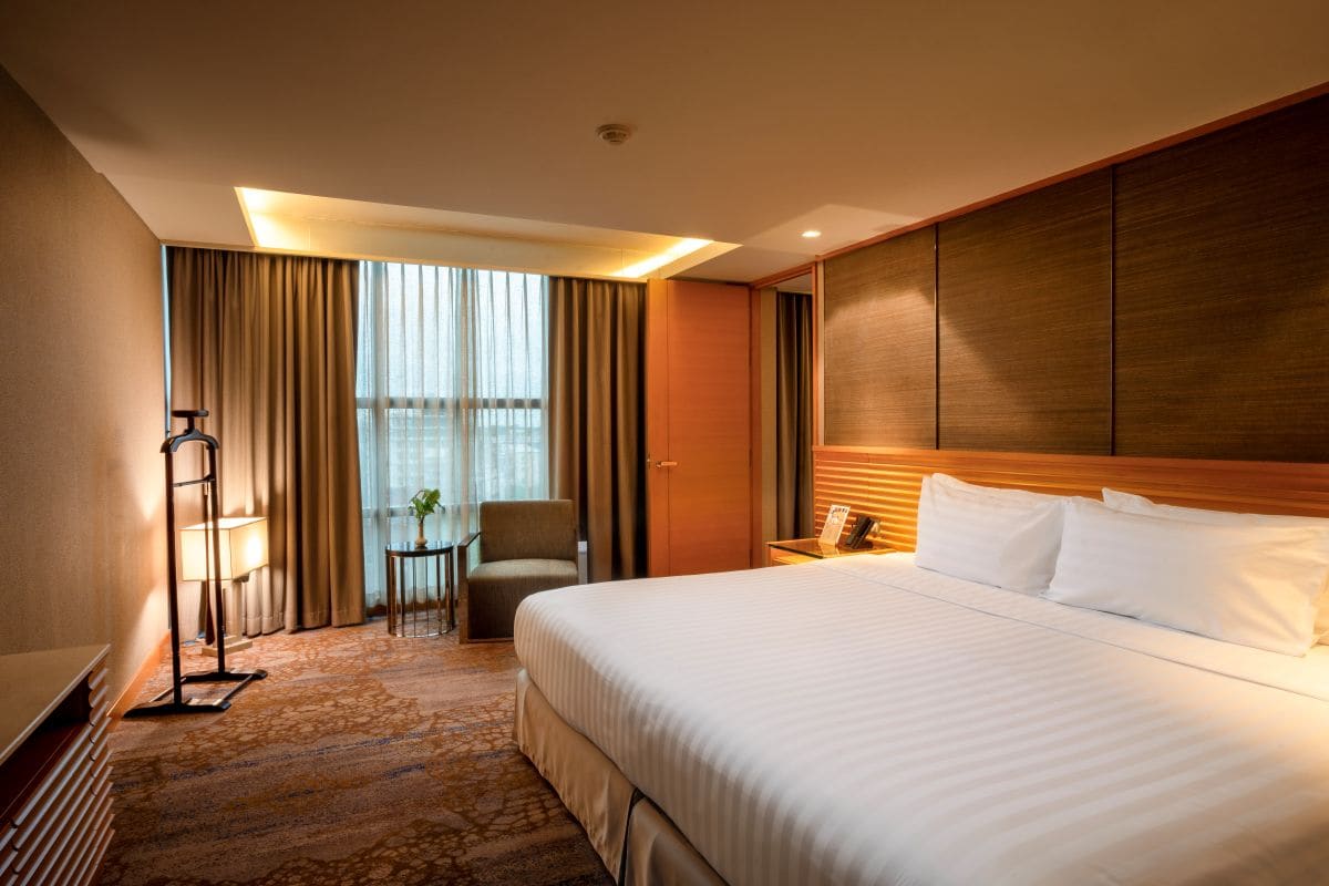 Quarto de hotel com uma cama grande, uma escrivaninha, uma cadeira, uma mesinha e vista para o exterior pela janela.