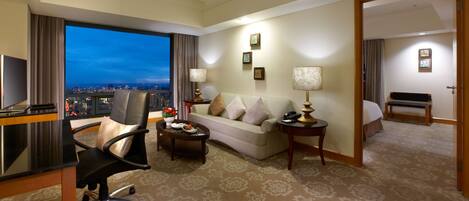 A hotel room with a city view, a sofa, a desk, a chair, a lamp, and a bedside table.
