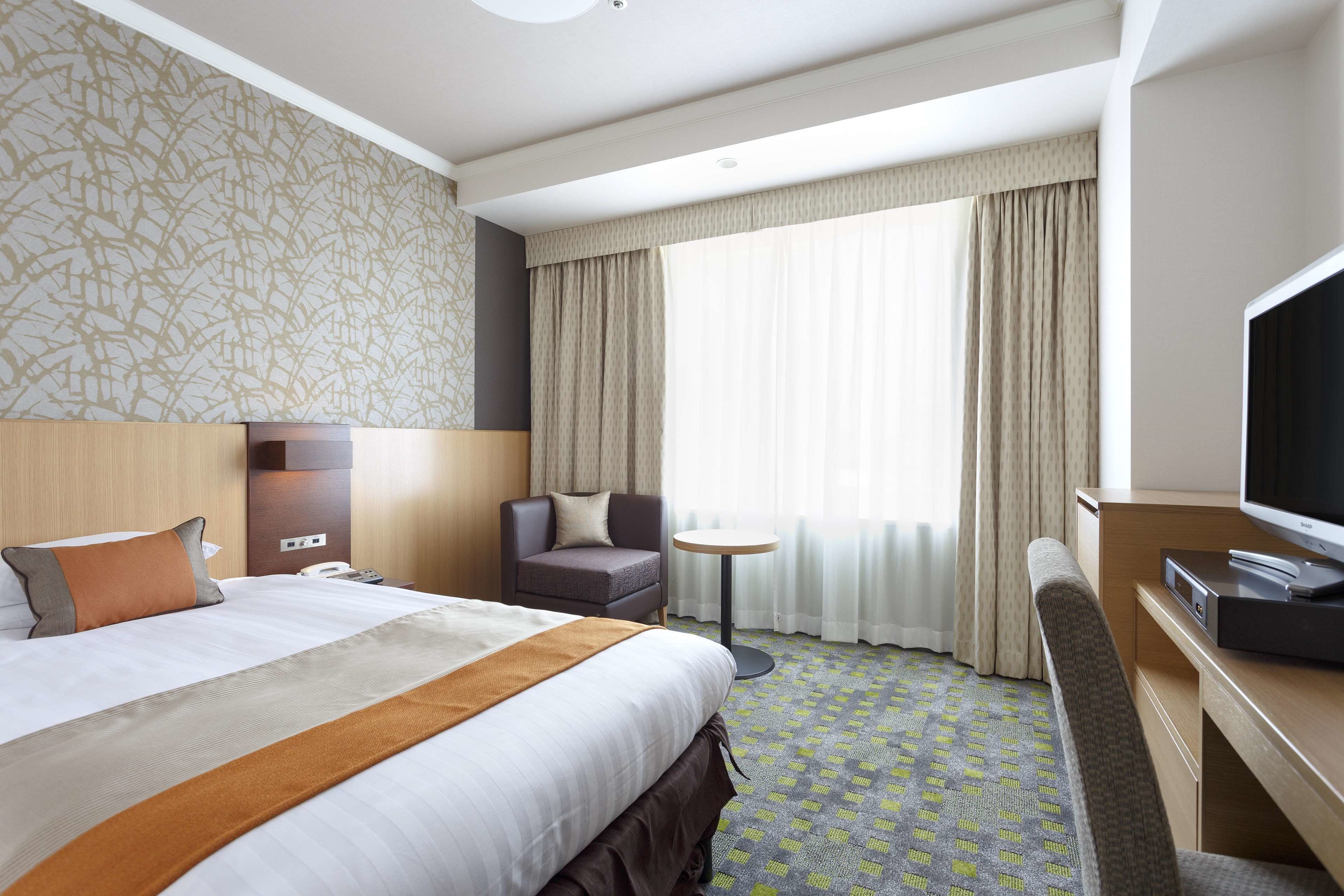 A hotel room with a large bed, a desk with a television, a chair, a small round table, and a window with curtains.