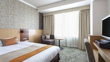 A hotel room with a large bed, a desk with a television, a chair, a small round table, and a window with curtains.