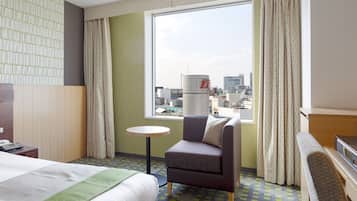 Habitación de hotel con cama, escritorio con teléfono, silla, mesita redonda, televisor y vista de la ciudad a través de un ventanal grande.