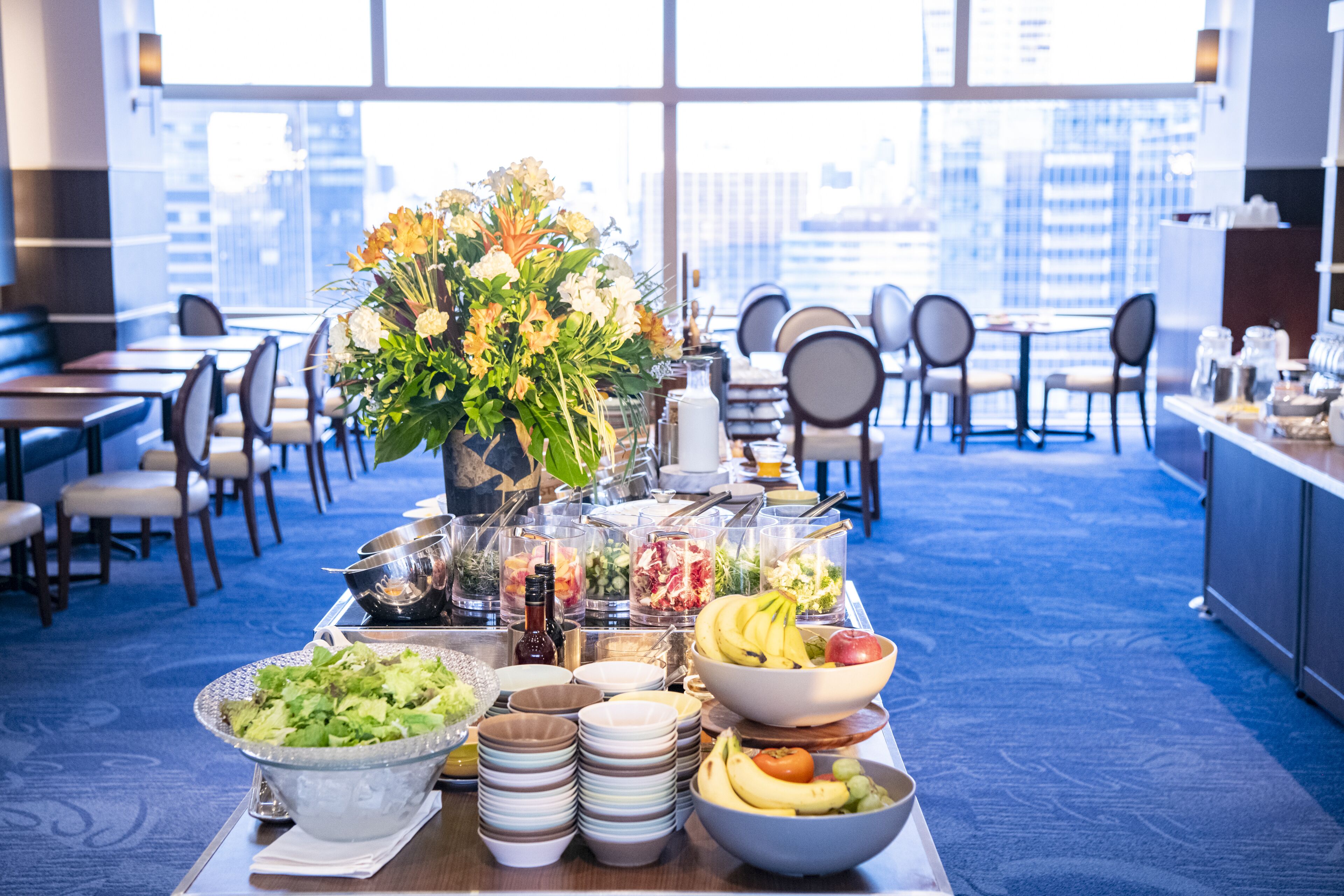 Un buffet proposant une variété de plats, agrémenté d’un arrangement floral en guise de centre de table, dans un espace de restauration moderne offrant une vue sur la ville.