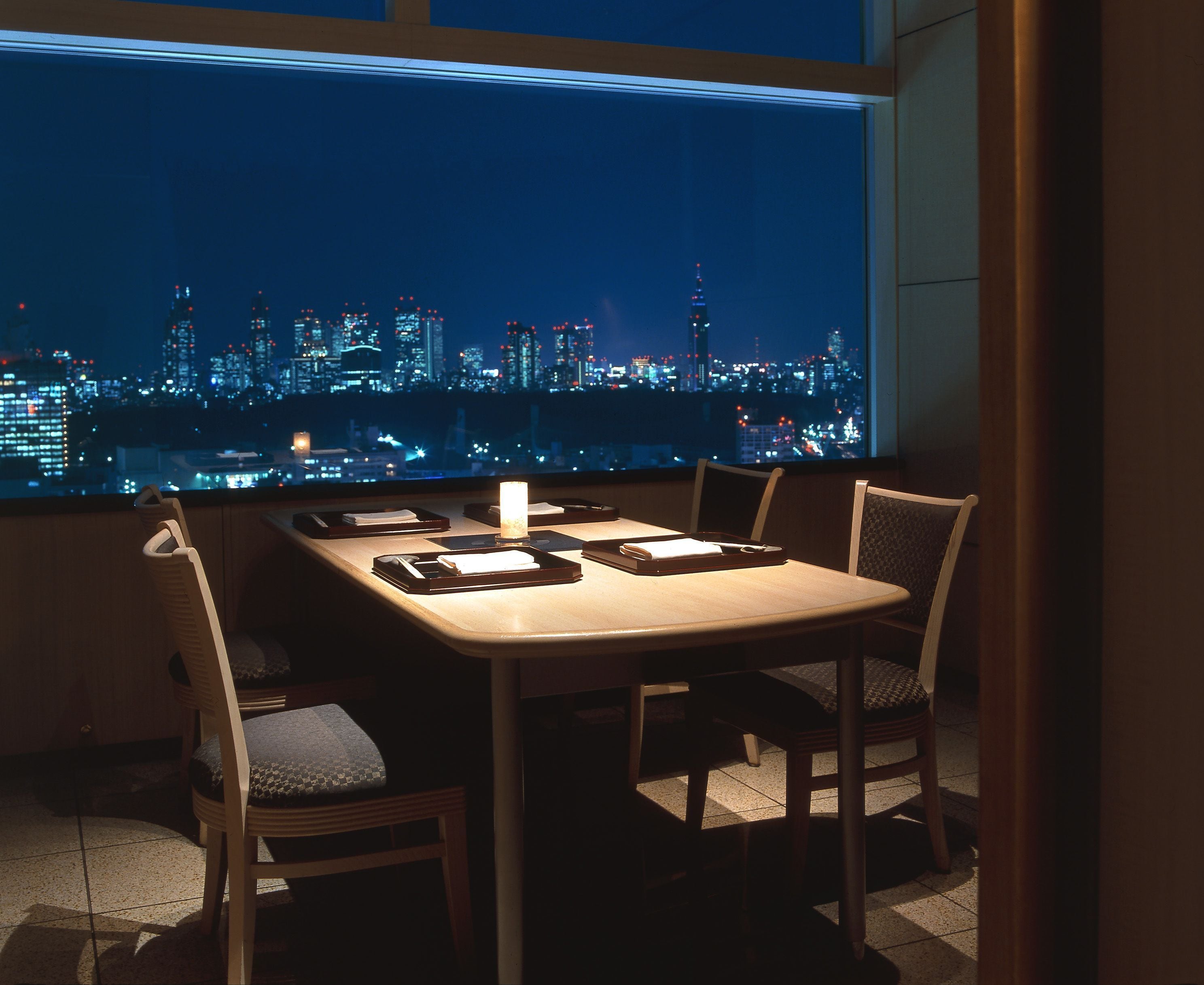 A dining area with a table set for four, chairs, and a candle. A cityscape is visible through a large window.