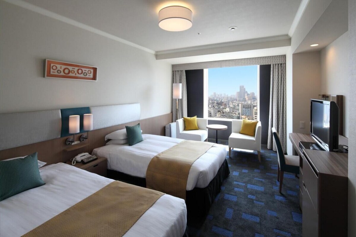 A hotel room with two beds, a TV, a desk, and a city view.
