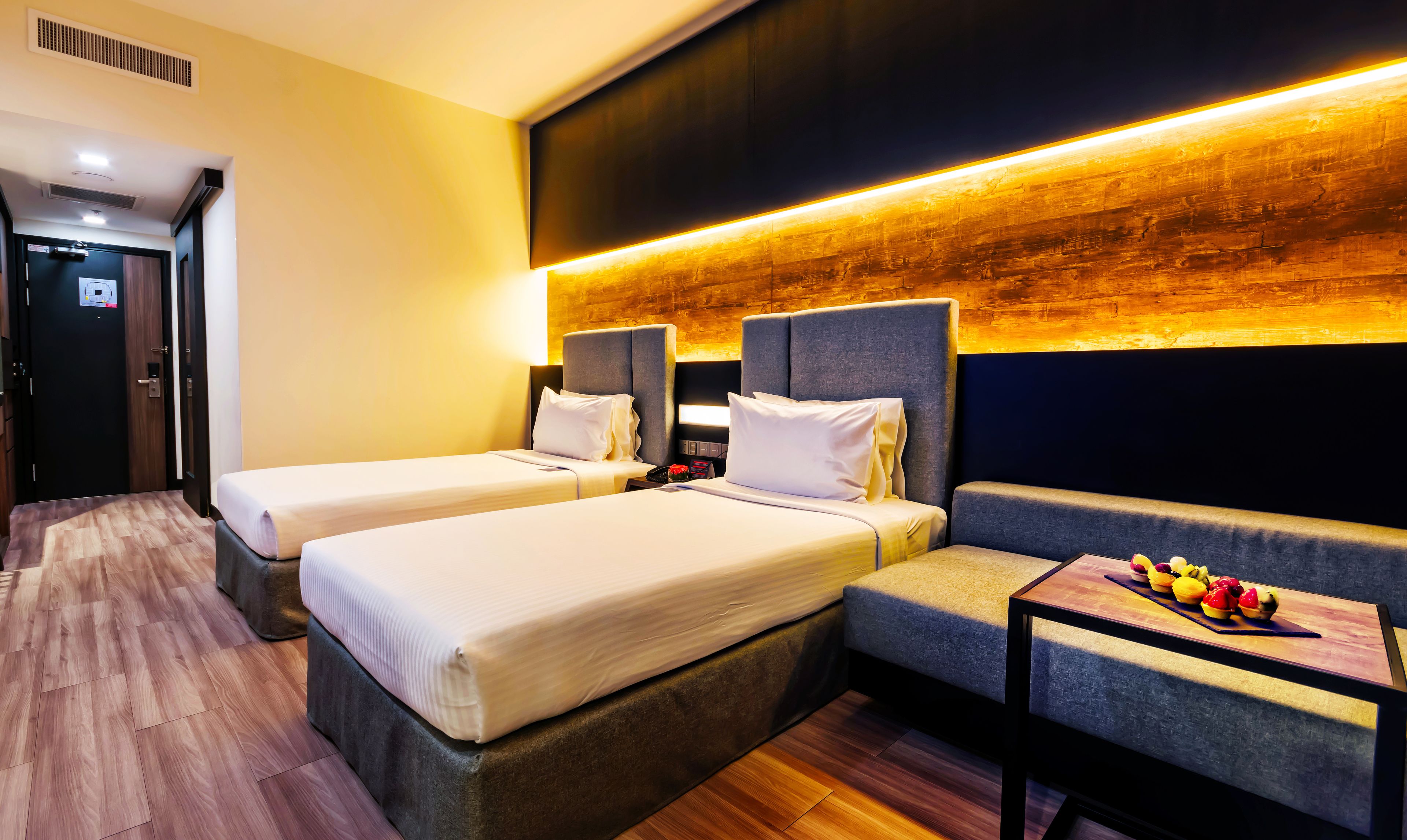 Quarto de hotel com duas camas individuais, um sofá e uma mesinha com frutas.