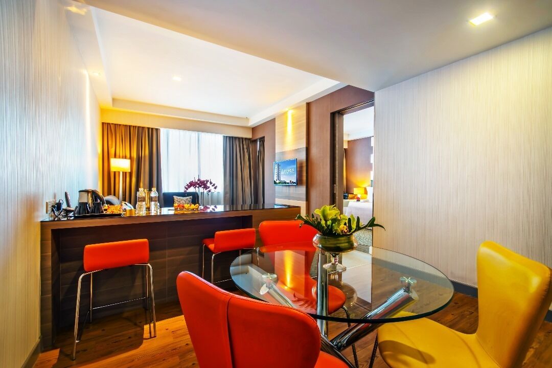 A modern hotel room with a glass dining table, red chairs, a bar area, and a television.