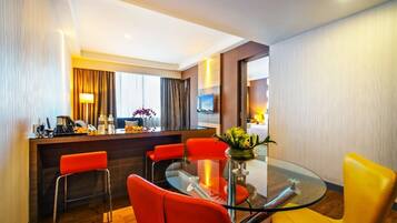 A modern hotel room with a glass dining table, red chairs, a bar area, and a television.
