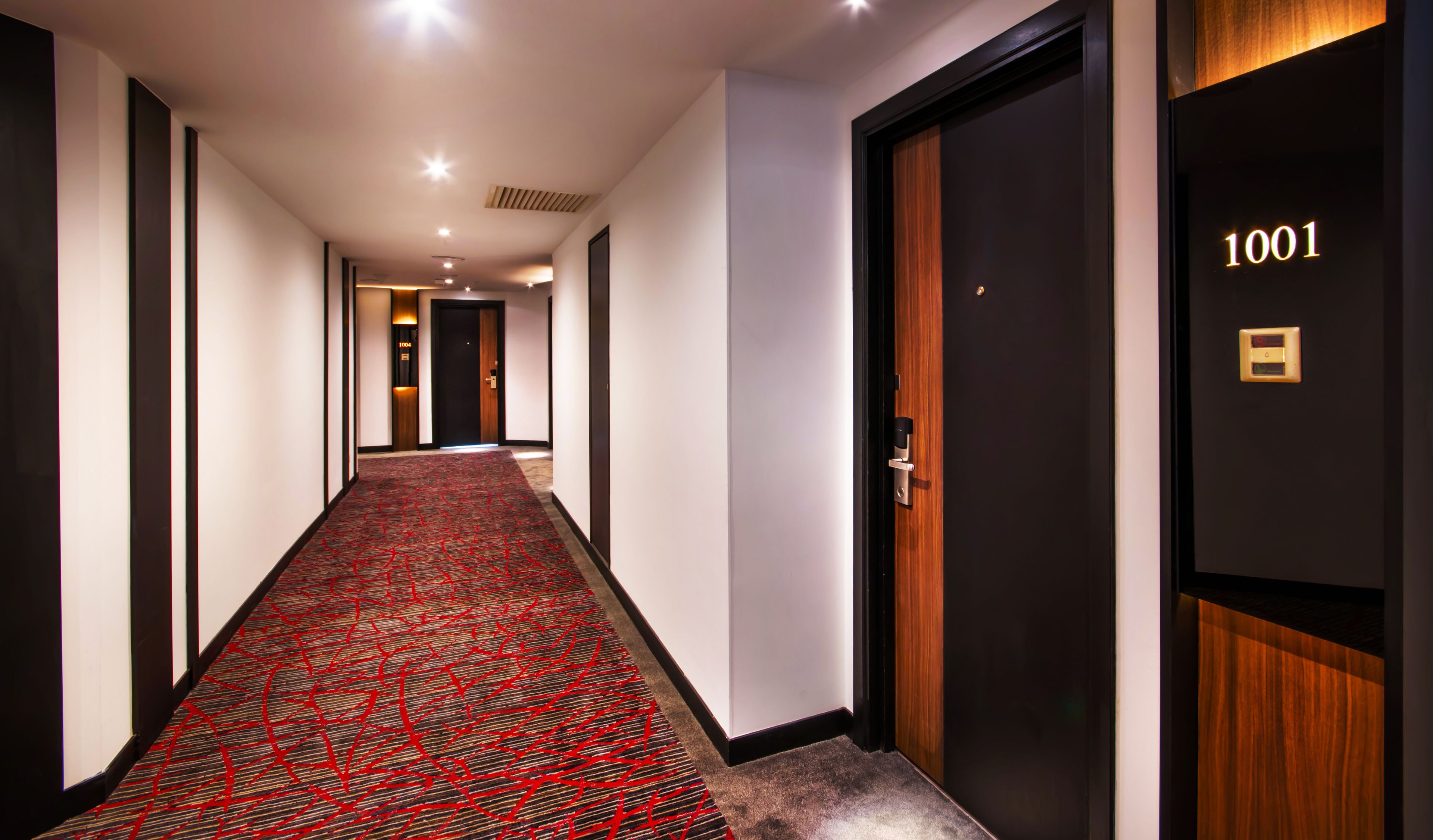 Un pasillo de hotel con una alfombra roja estampada, puertas de madera oscura y el número 1001 en una de ellas.