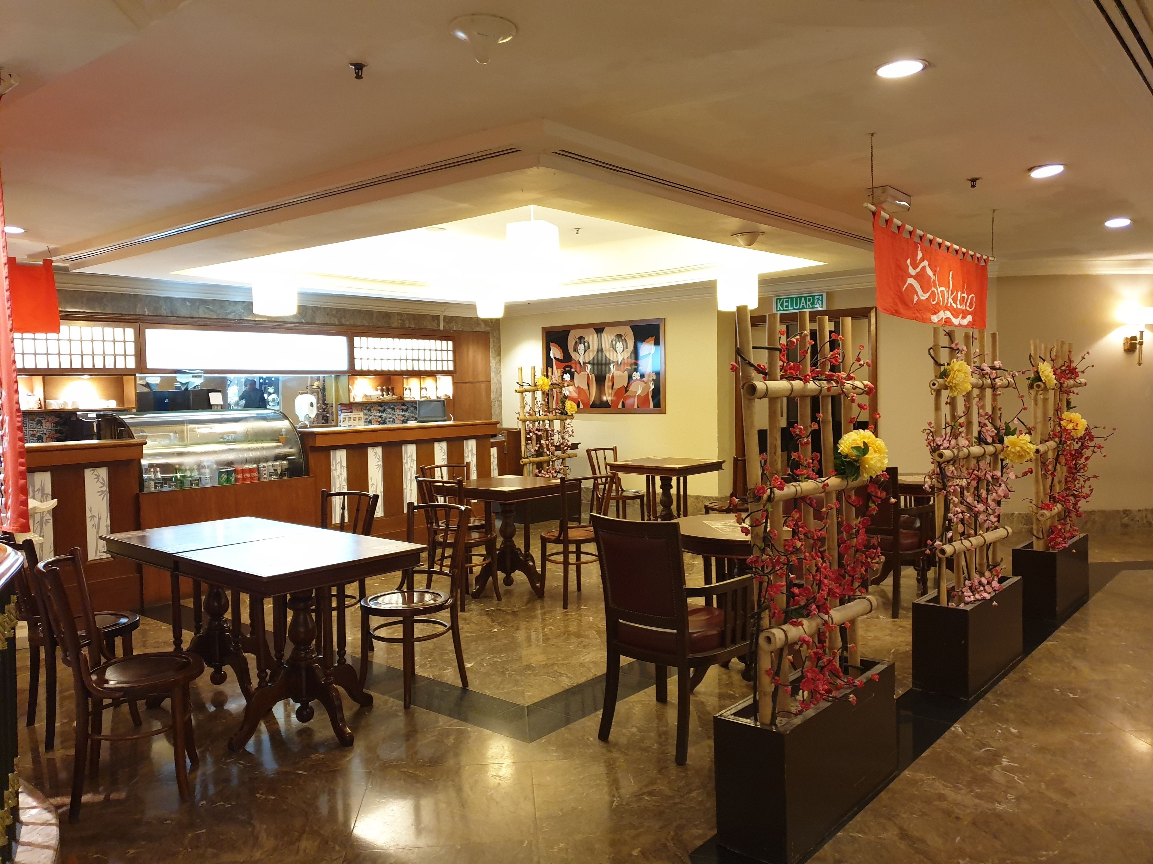 Un interior de restaurante con mesas y sillas de madera, una barra con vitrina y arreglos florales decorativos.