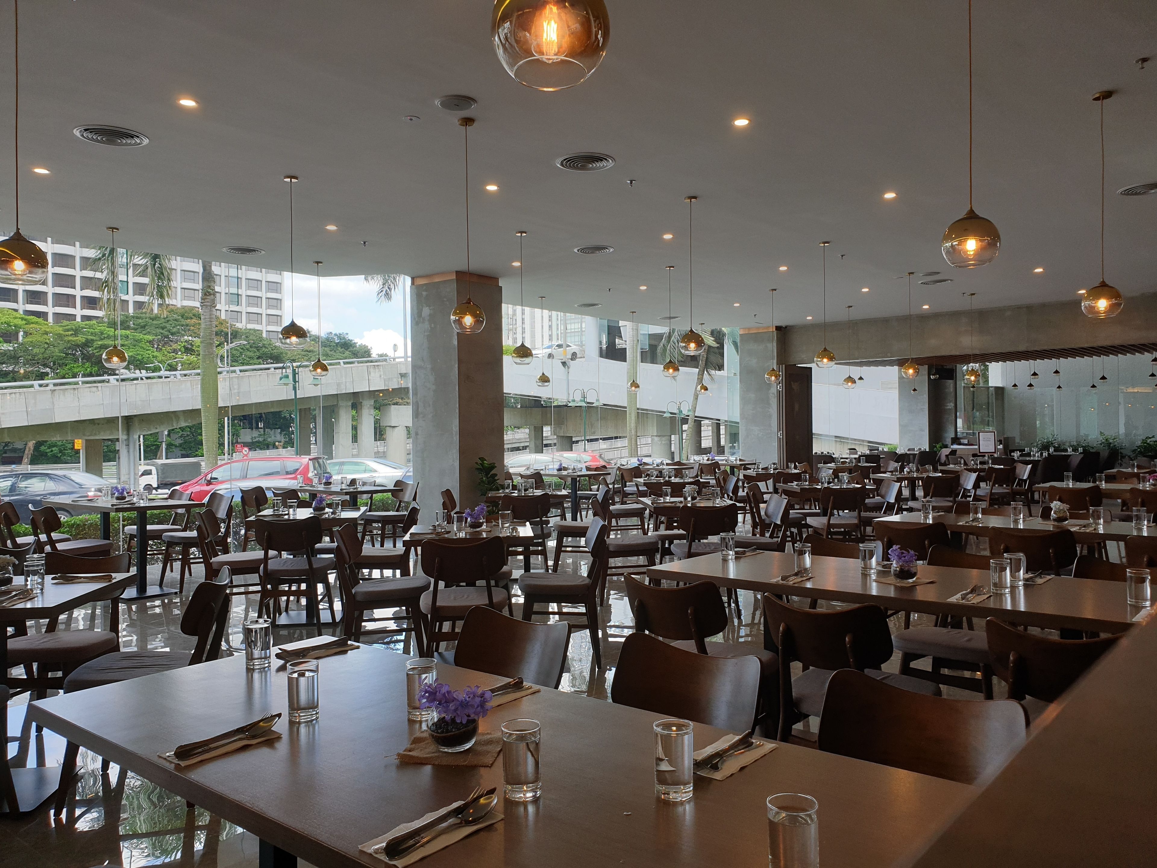 A spacious restaurant with multiple tables and chairs, large windows offering a view of the street, and hanging pendant lights.