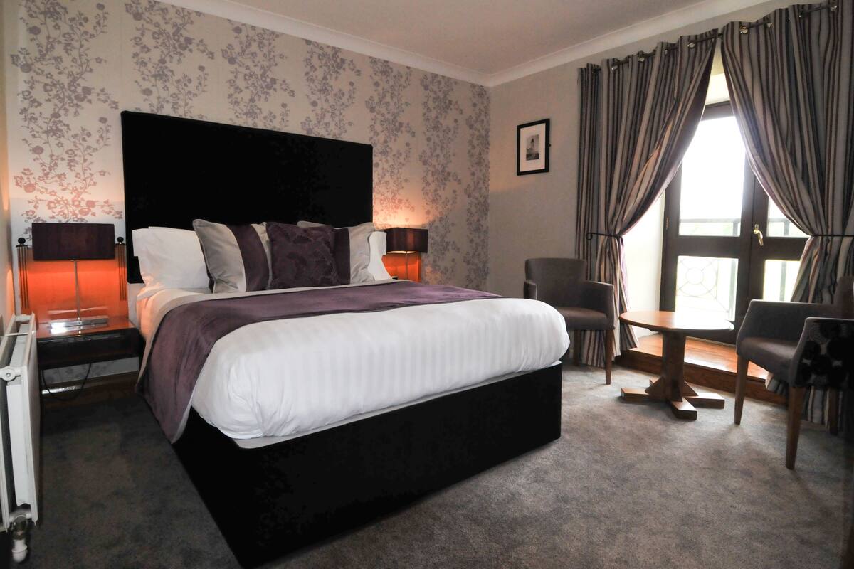 A hotel room with a large bed, two bedside tables with lamps, a chair, a small table, and a window with curtains.