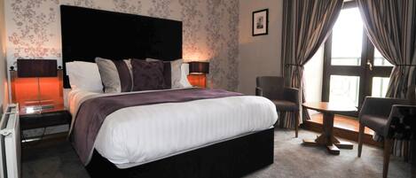 A hotel room with a large bed, two bedside tables with lamps, a chair, a small table, and a window with curtains.