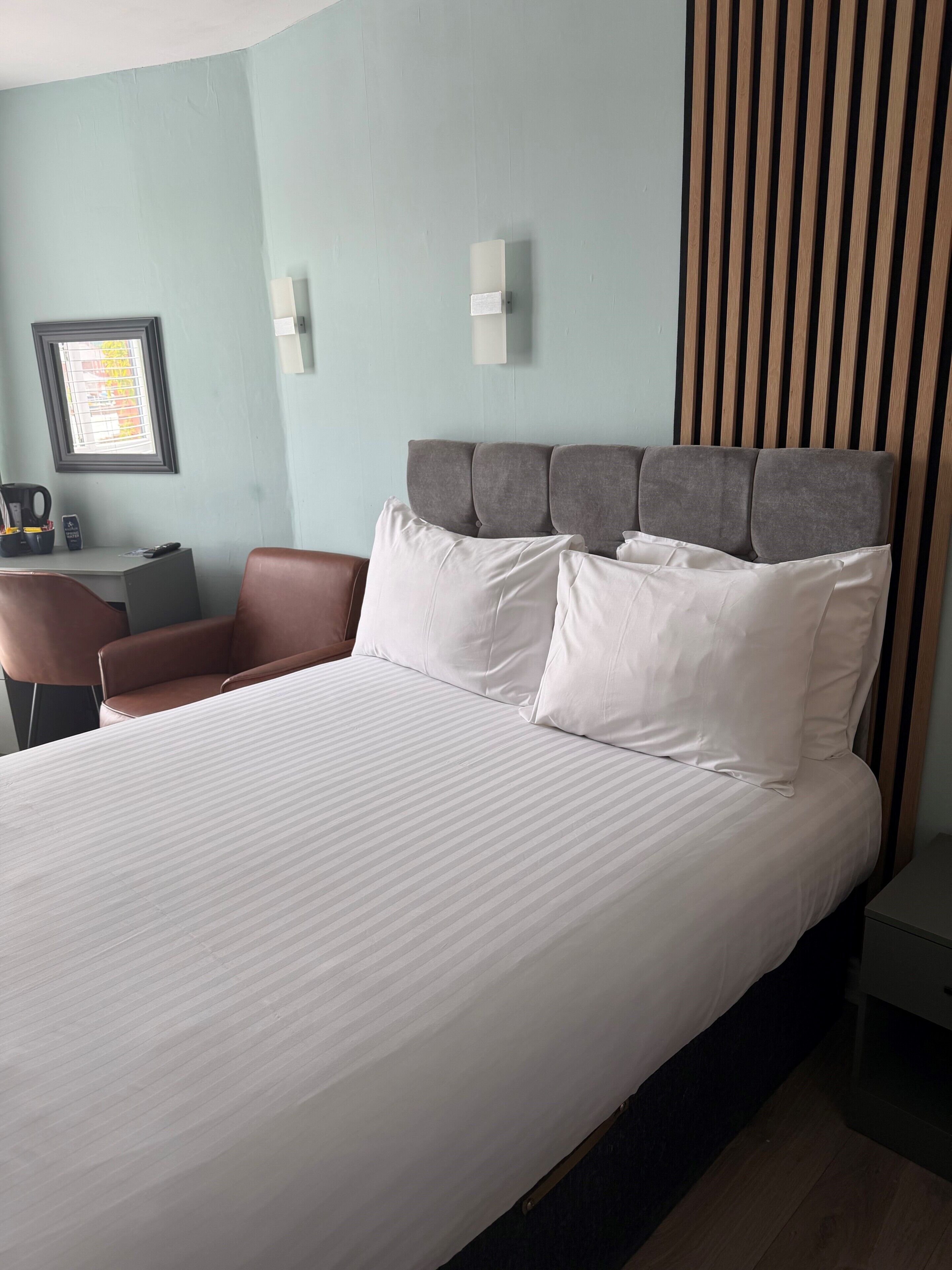 A hotel room with a large bed, a desk with a chair, a window with blinds, and a wall-mounted light fixture.