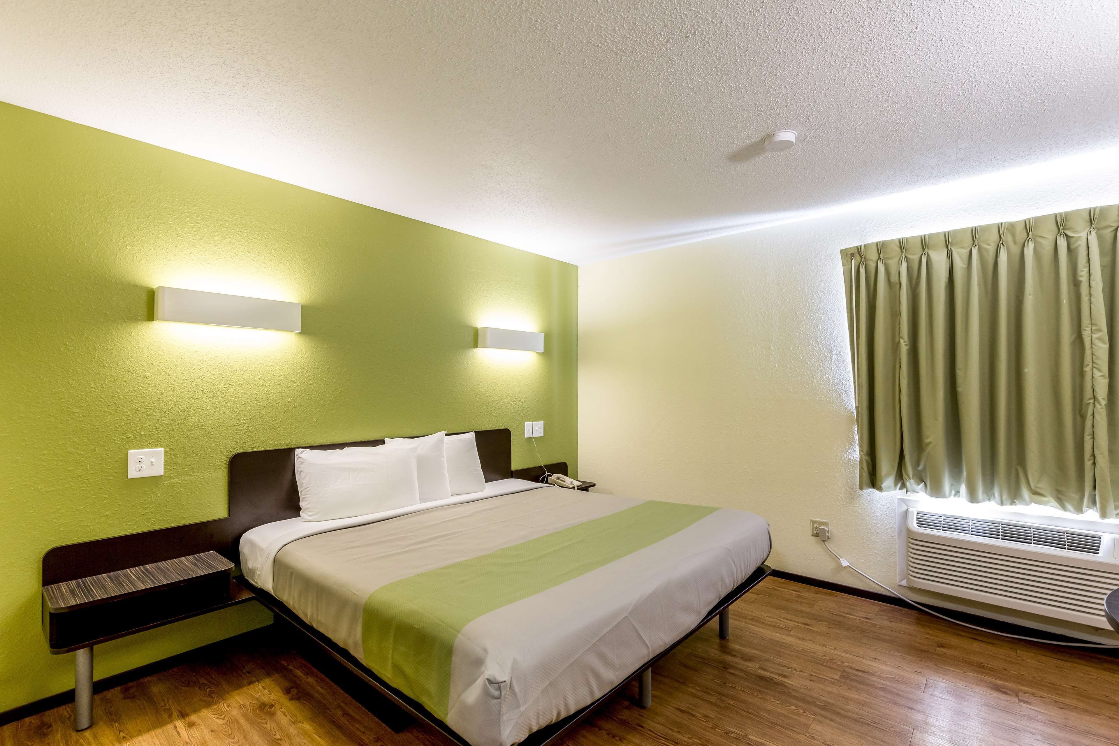 A hotel room with a bed, bedside table, wall-mounted light, air conditioning unit, and a window with curtains.