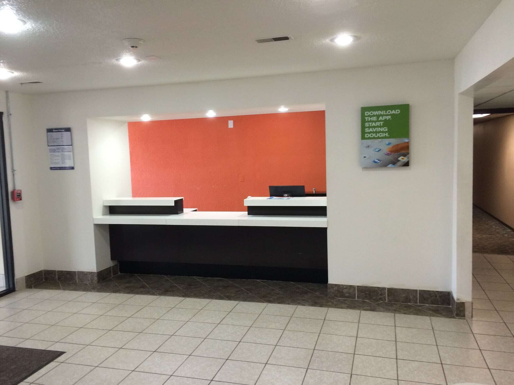 Un comptoir d’accueil avec un plan de travail blanc et un mur orange, une affiche faisant la promotion d’une application et un sol carrelé.