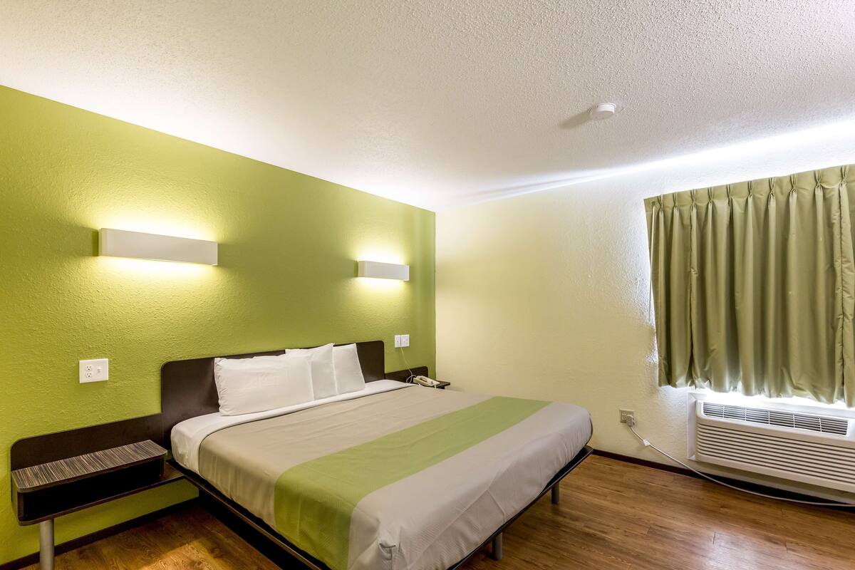 A hotel room with a bed, bedside table, wall-mounted light, air conditioning unit, and a window with curtains.