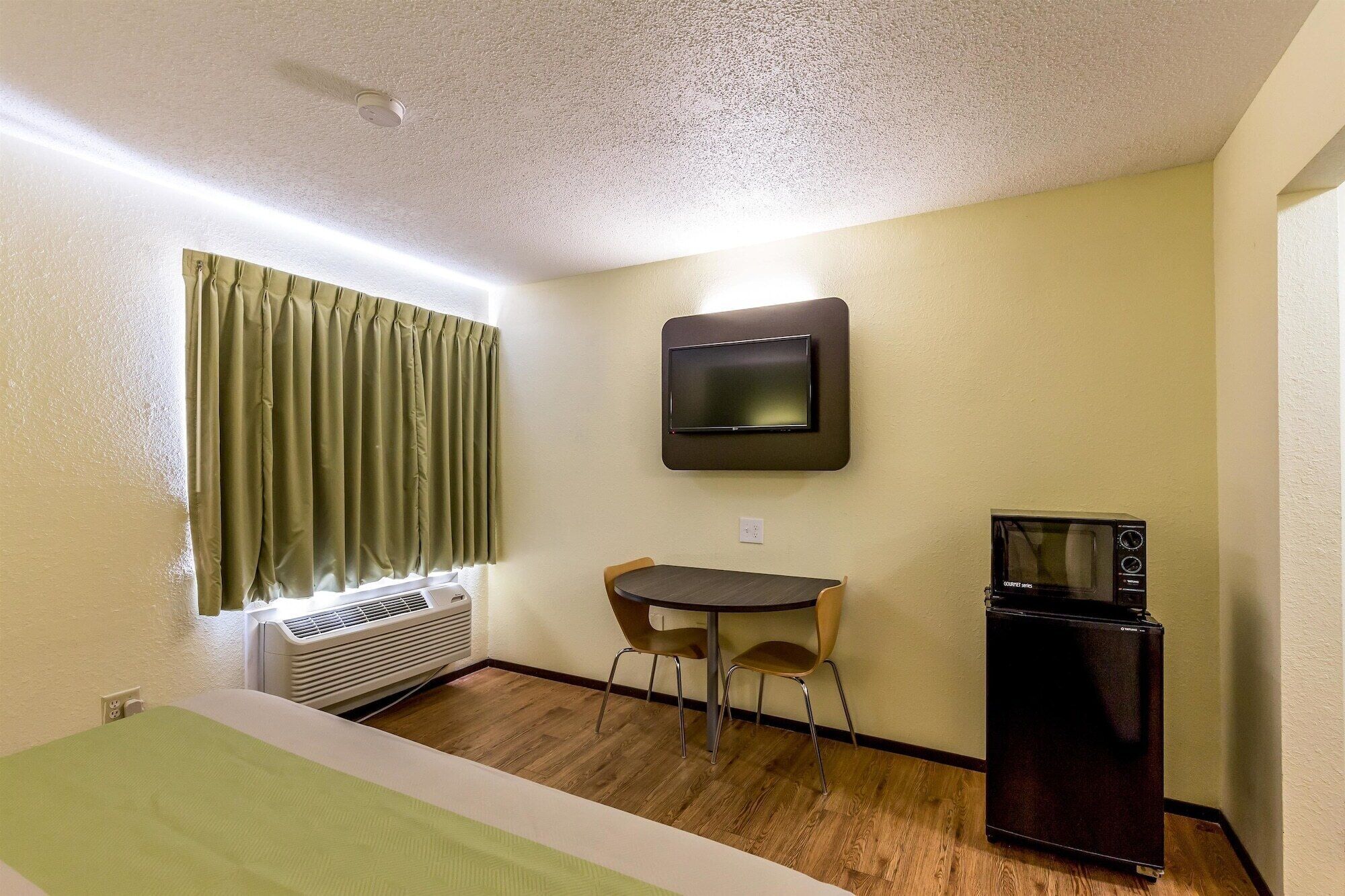 A small room with a bed, a table with two chairs, a TV mounted on the wall, and a refrigerator.