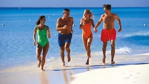 Quatre personnes courent sur une plage, avec une eau d’un bleu limpide et une mer calme en arrière-plan.