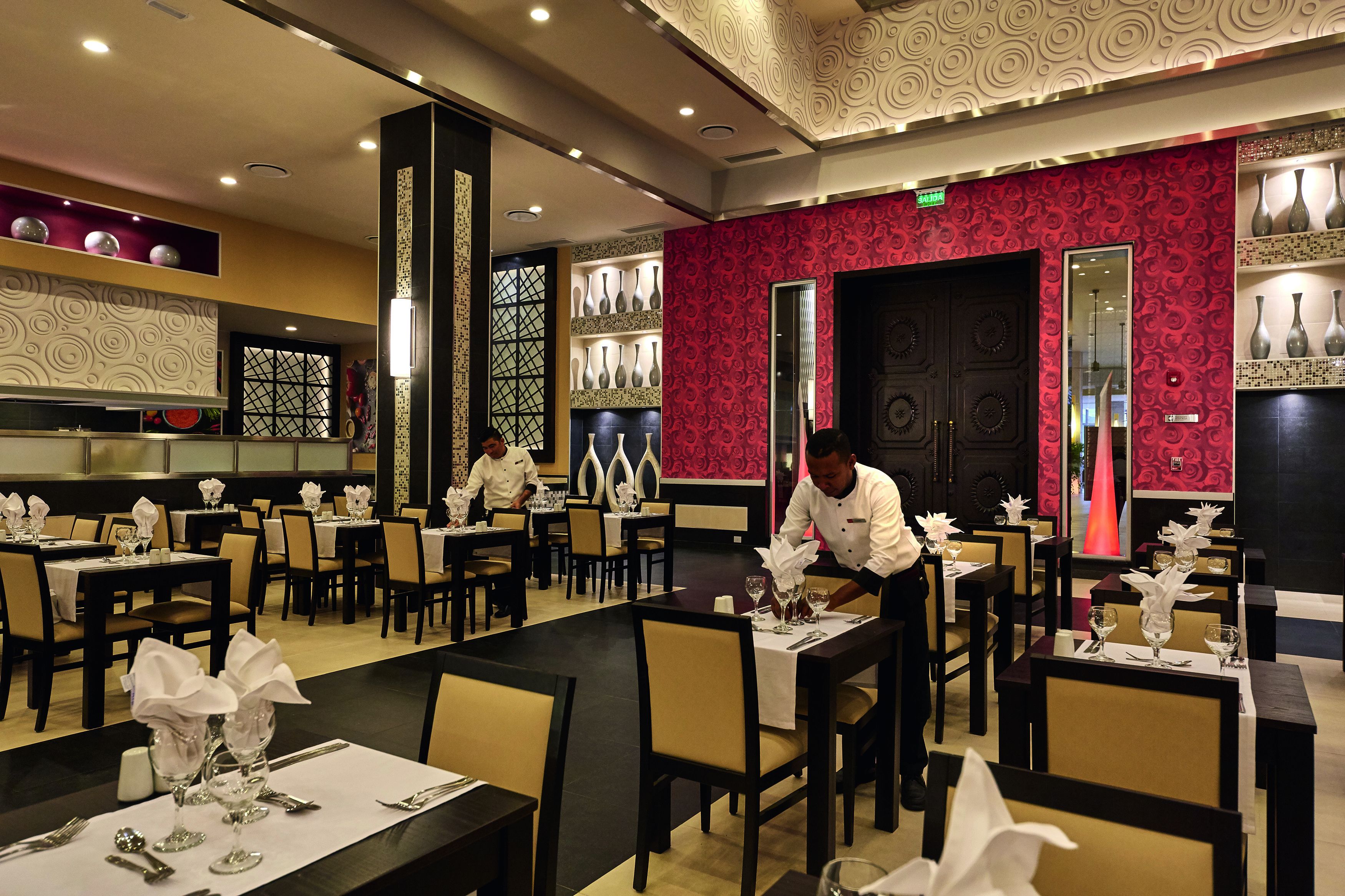 Un restaurant avec plusieurs tables et chaises, un chef qui prépare les plats et des vases décoratifs disposés sur des étagères.