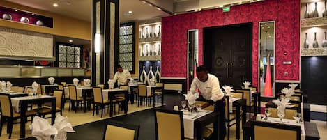 A restaurant with multiple tables and chairs, a chef preparing food, and decorative vases on shelves.