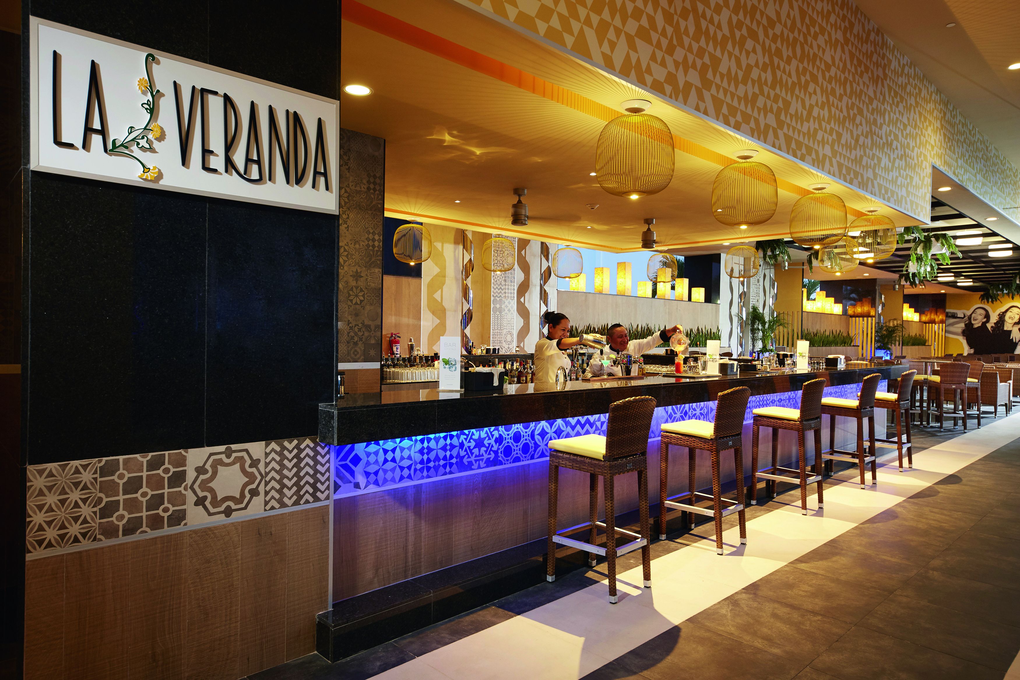 A bar area with a sign reading "LA VERANDA," featuring a counter with stools, a patterned wall, and hanging lights.