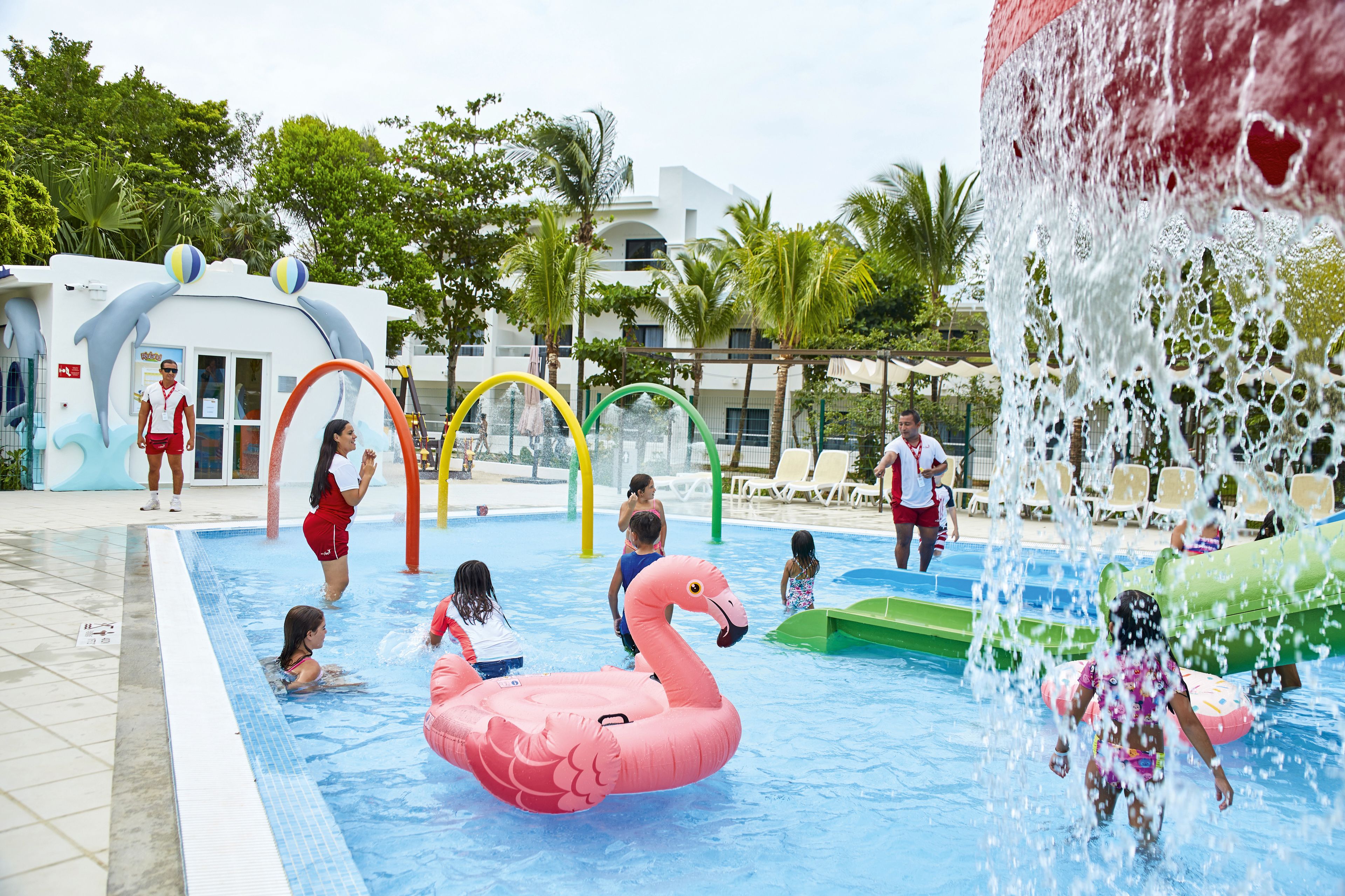Une piscine où des enfants jouent, un flotteur en forme de flamant rose et une fontaine.
