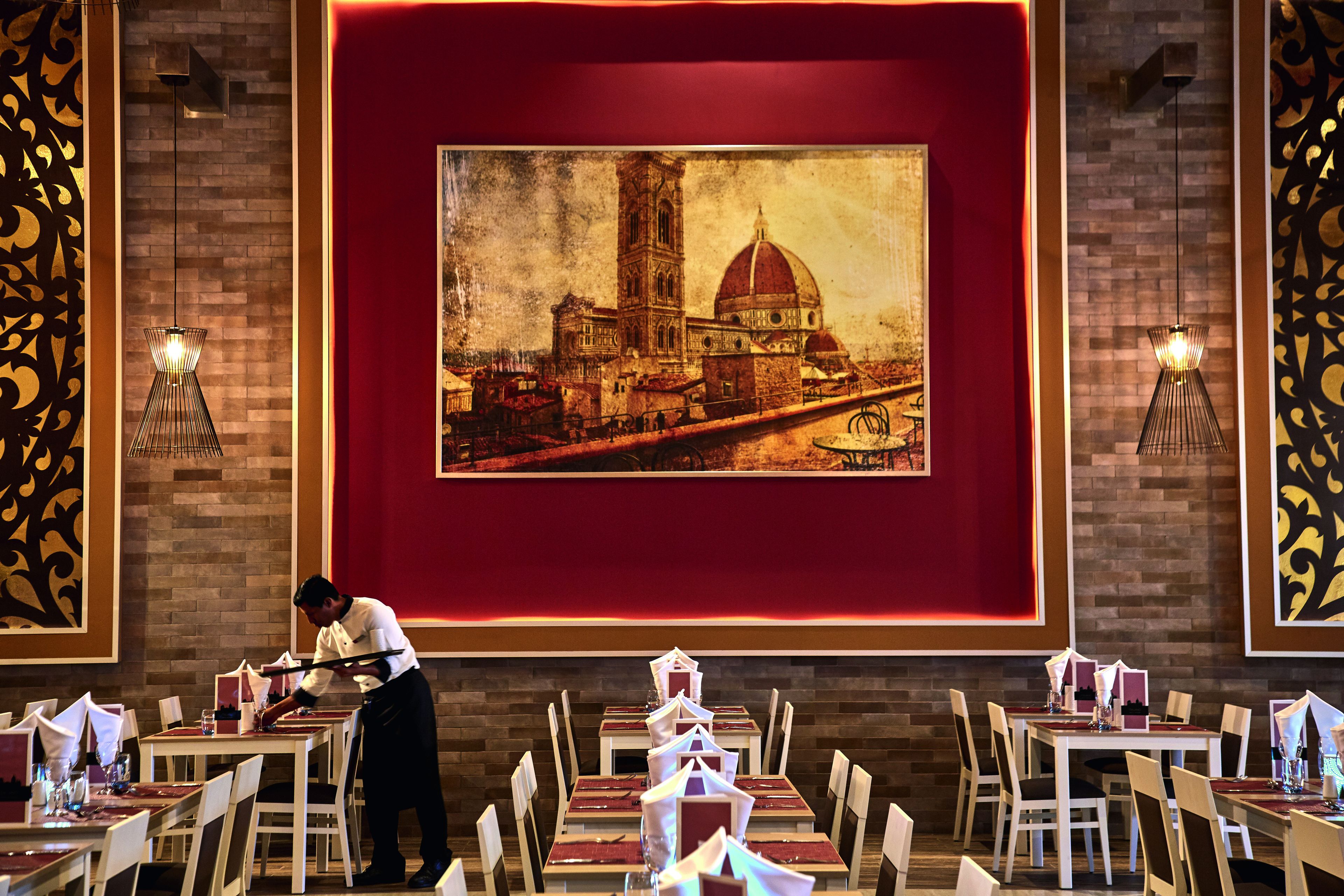 Un restaurant où un serveur prépare les tables. Un grand tableau représentant un bâtiment historique est accroché au mur.