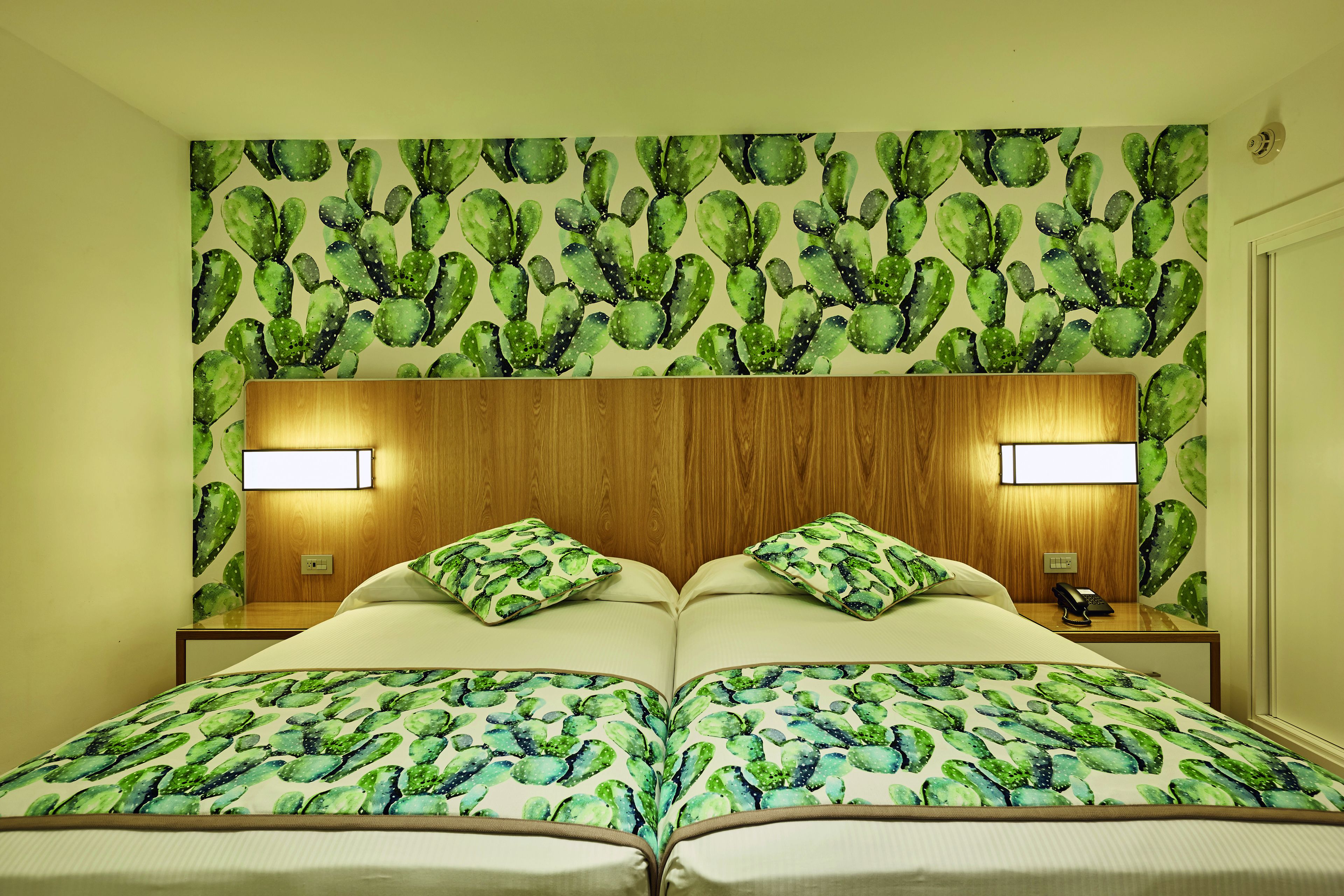 Un lit double avec une couvre-lit à motifs de cactus verts, deux lampes de chevet et une tête de lit en bois.