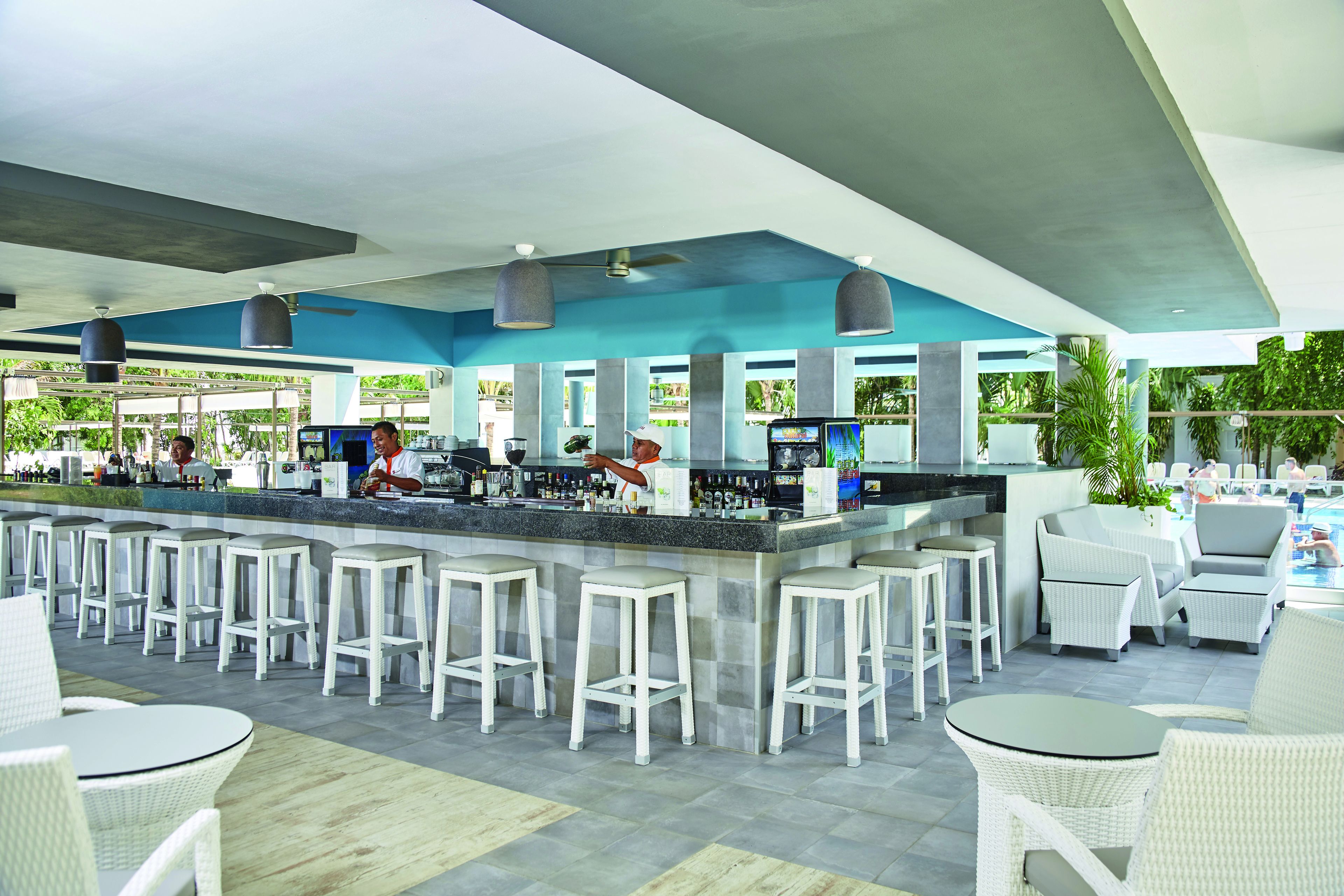A bar area with white bar stools, a counter with a bartender, and outdoor seating with white wicker furniture.