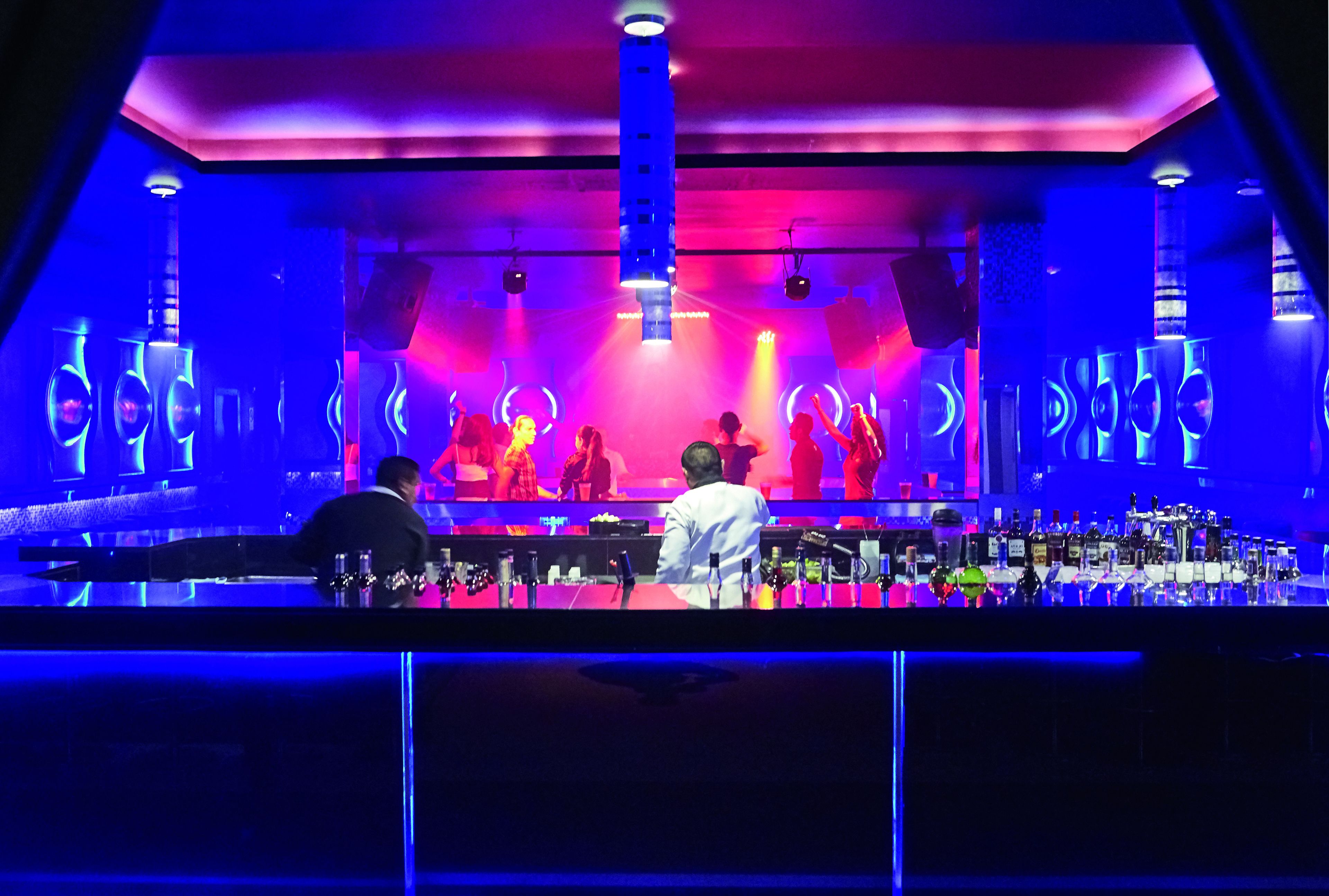 A bar area with a stage and DJ booth, illuminated by blue and pink lighting.