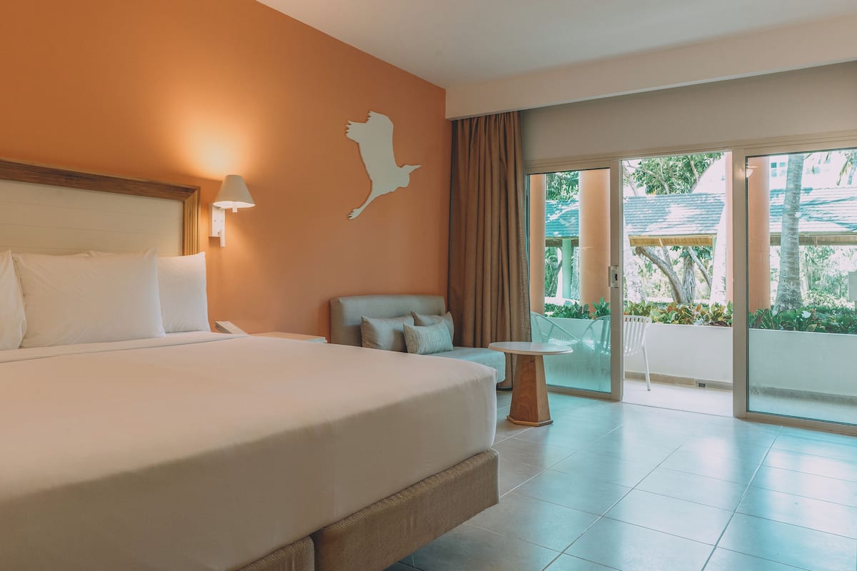 A hotel room with a large bed, a sofa, a small table, and a view of the outdoors through a sliding door.
