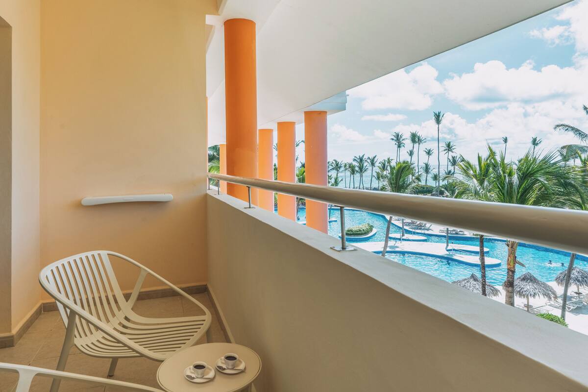 A balcony with a view of a swimming pool, palm trees, and orange pillars.