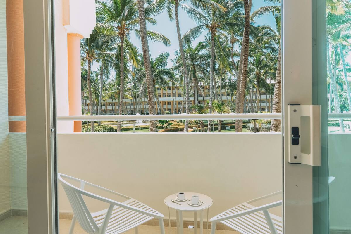 A balcony with white chairs and a small table, overlooking a palm tree-lined courtyard and a building with multiple balconies.